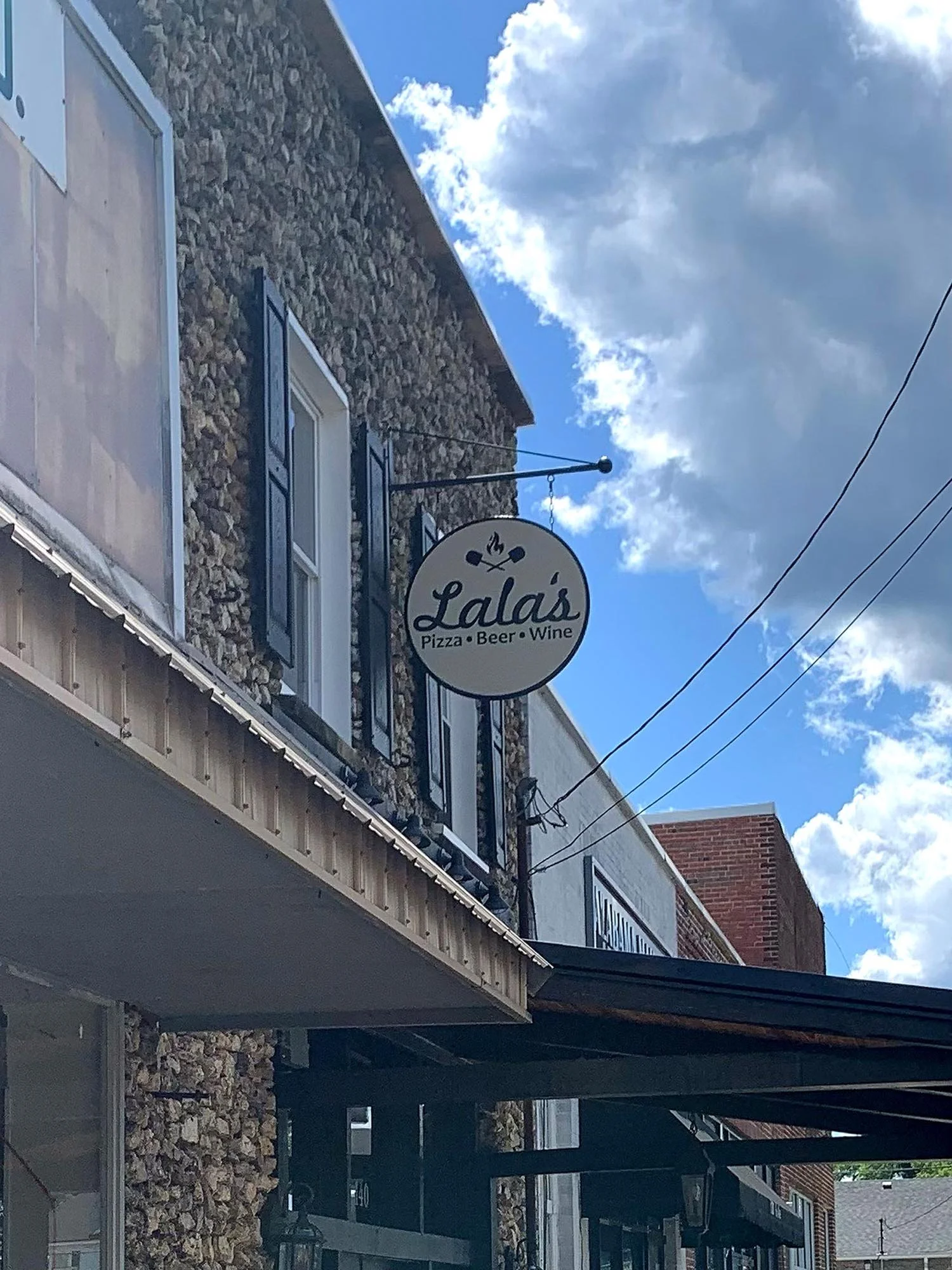 A sign for Lala's restaurant advertising pizza, beer, and wine, hanging outside on a building with a stone facade under a partly cloudy sky.