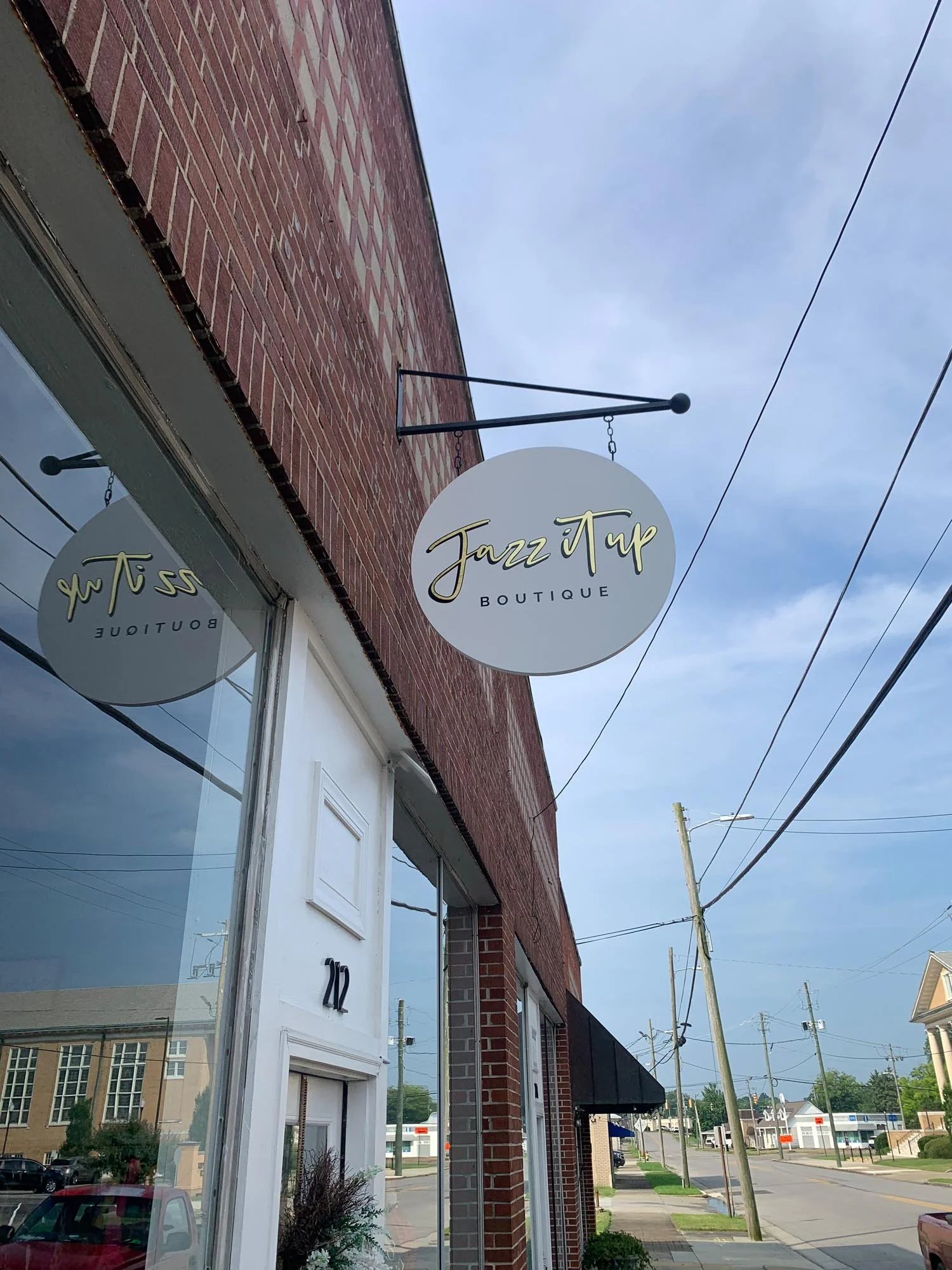 Exterior of a boutique store called Jazz it Up Boutique with a round sign hanging above the entrance, a brick building, a white door numbered 22, and power lines in the background on a cloudy day.