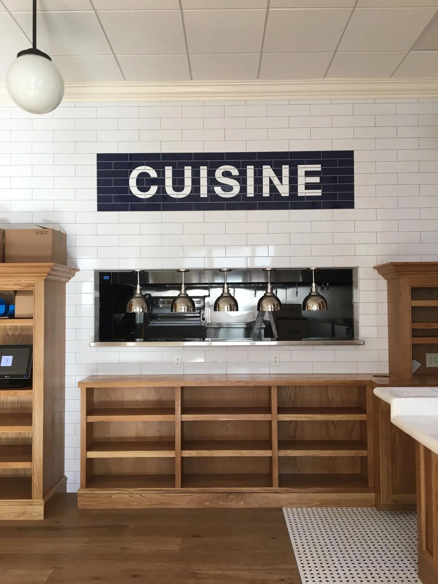 Interior of a restaurant with a white tiled wall and a black sign that says 'CUISINE'. There is a counter with stainless steel heat lamps over it and wooden shelves below. On the left side, there are additional wooden shelves and a small tablet or co