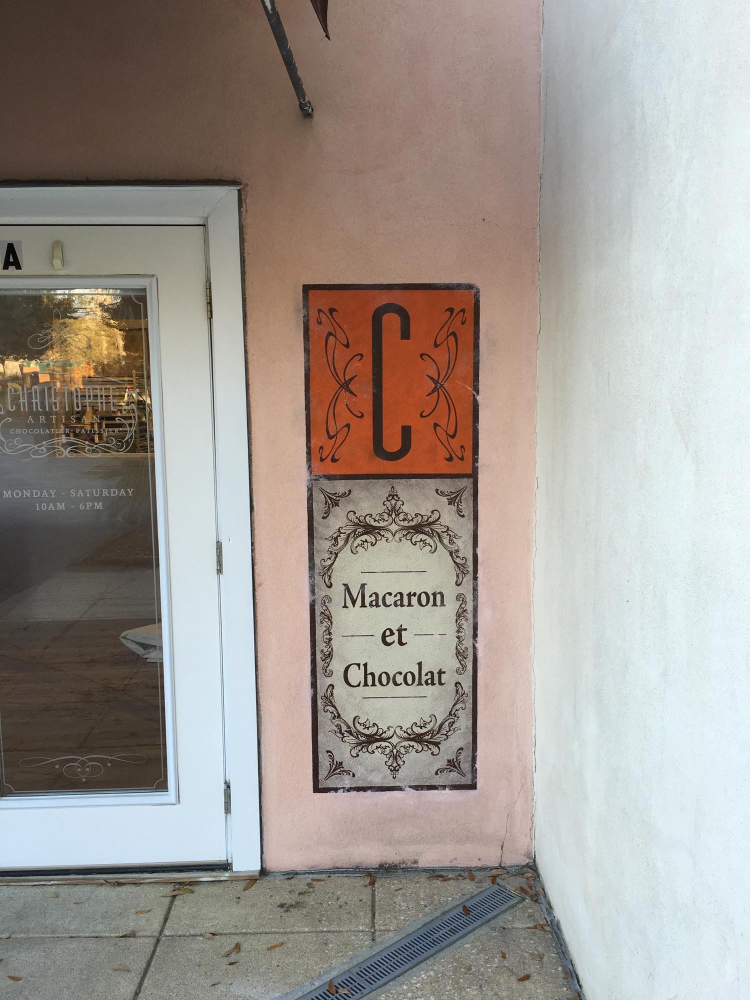 Painted sign outside a shop with decorative borders. The top part has a large letter 'C' on an orange background. The lower part reads 'Macaron et Chocolat' with ornate decorative borders.