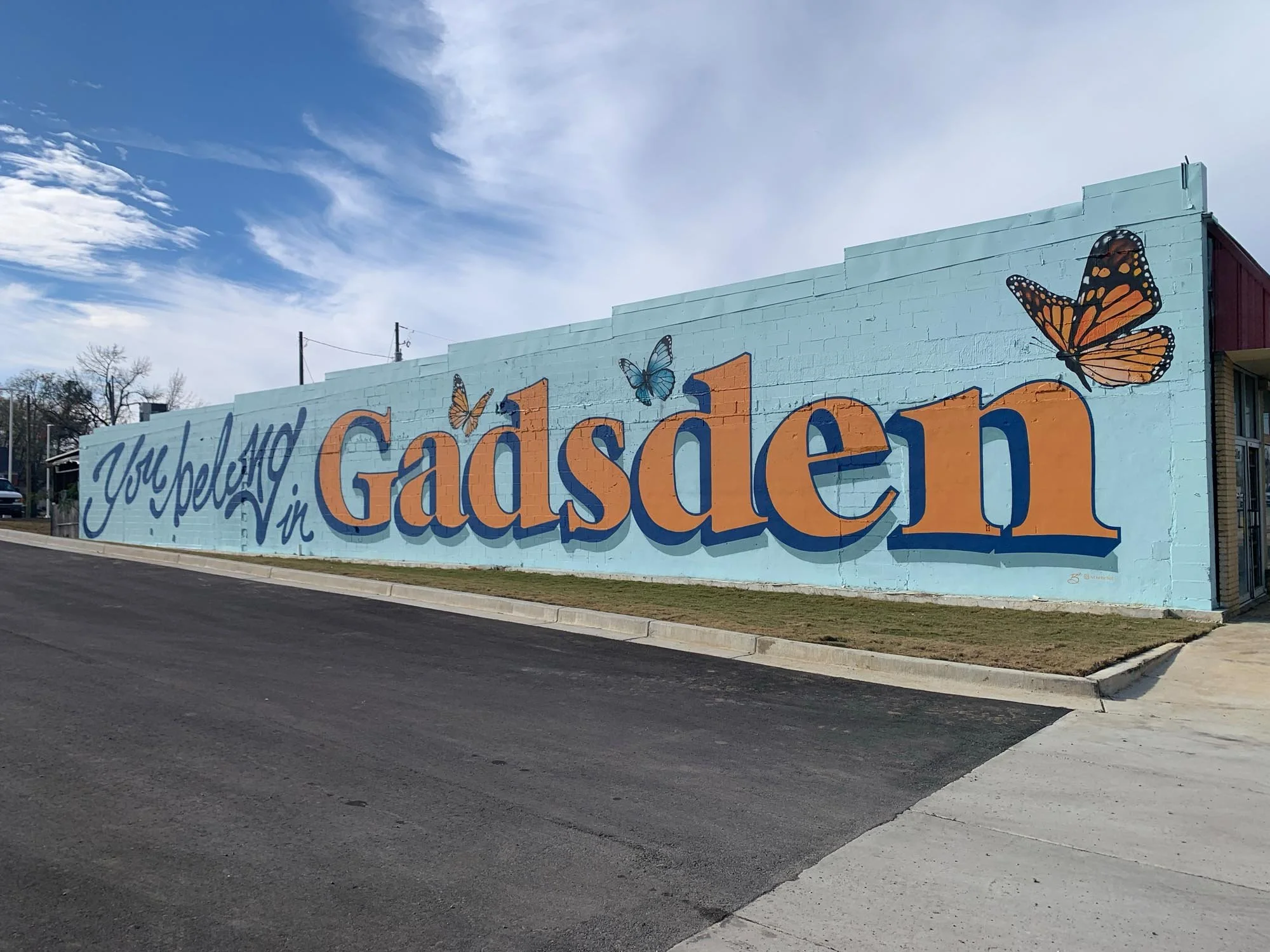 Colorful mural on a building wall that says 'You're helping in Gadsden' with butterflies and a blue sky.