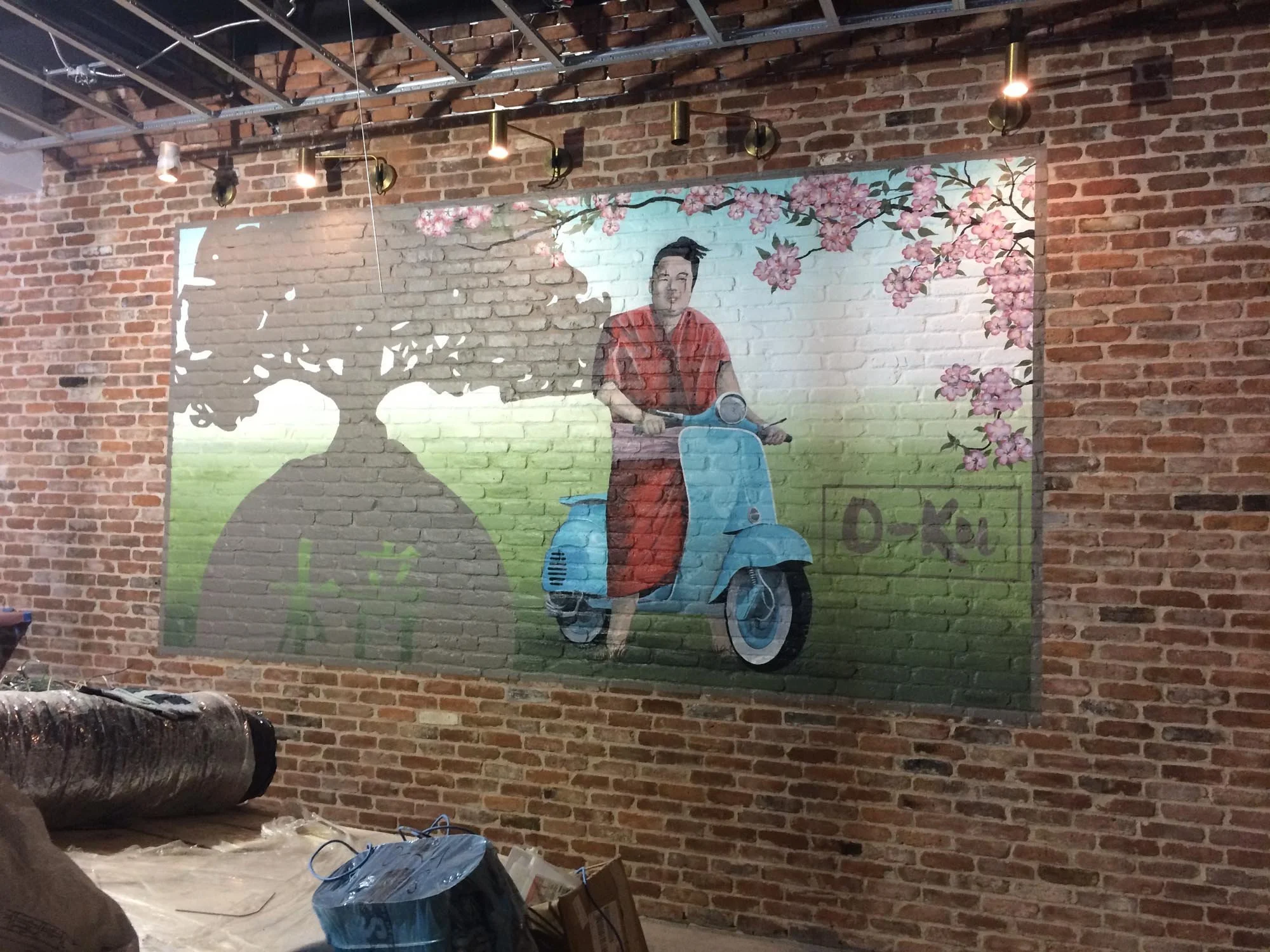 A mural depicting a woman in a red dress riding a light blue scooter with pink blossoms and a large tree shadow in the background, painted on an exposed brick wall.