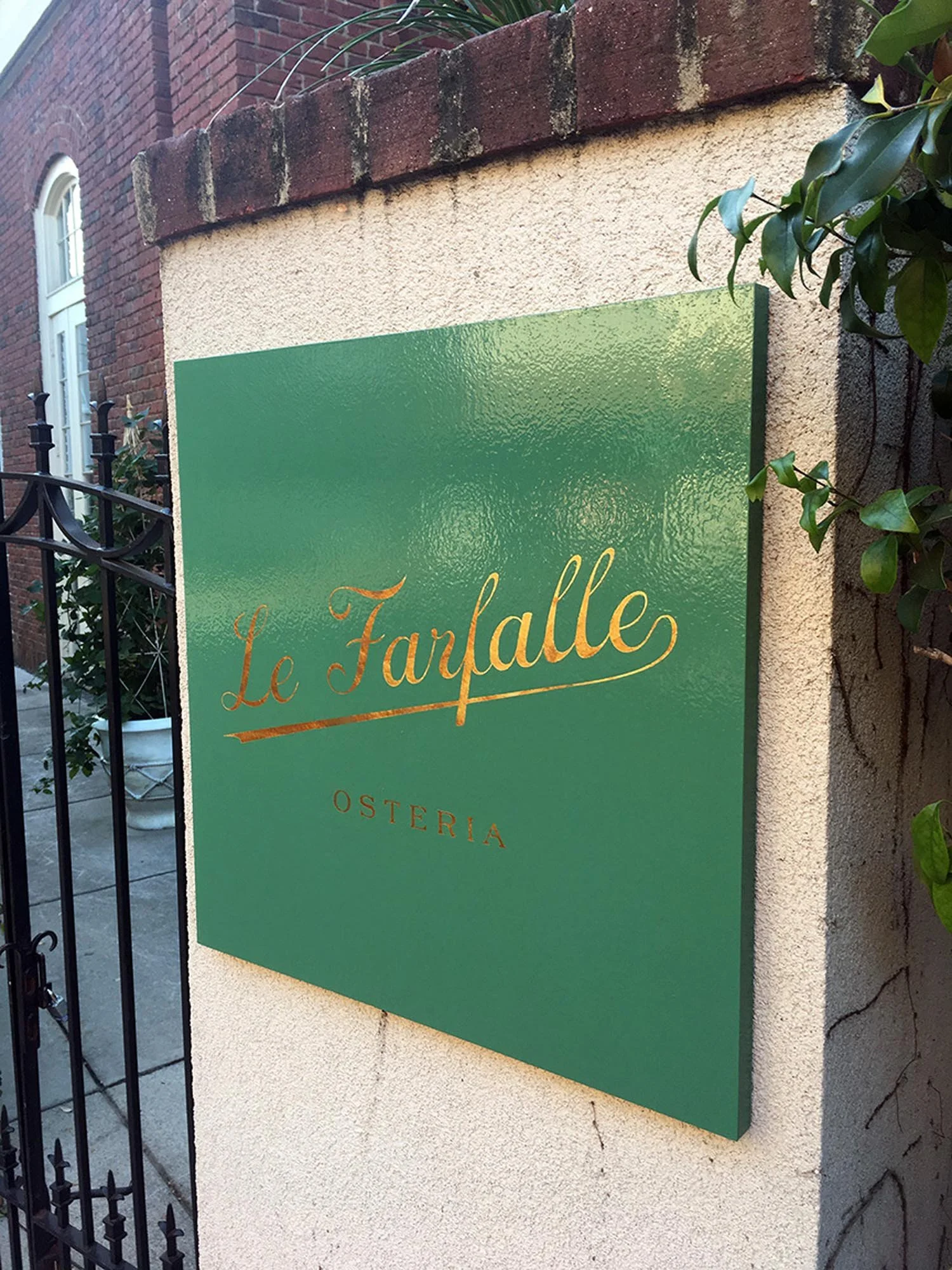 Green signboard with gold cursive text 'Le Fanfalle' and smaller text 'OSTERIA' underneath, mounted on a cream-colored wall outside a building.