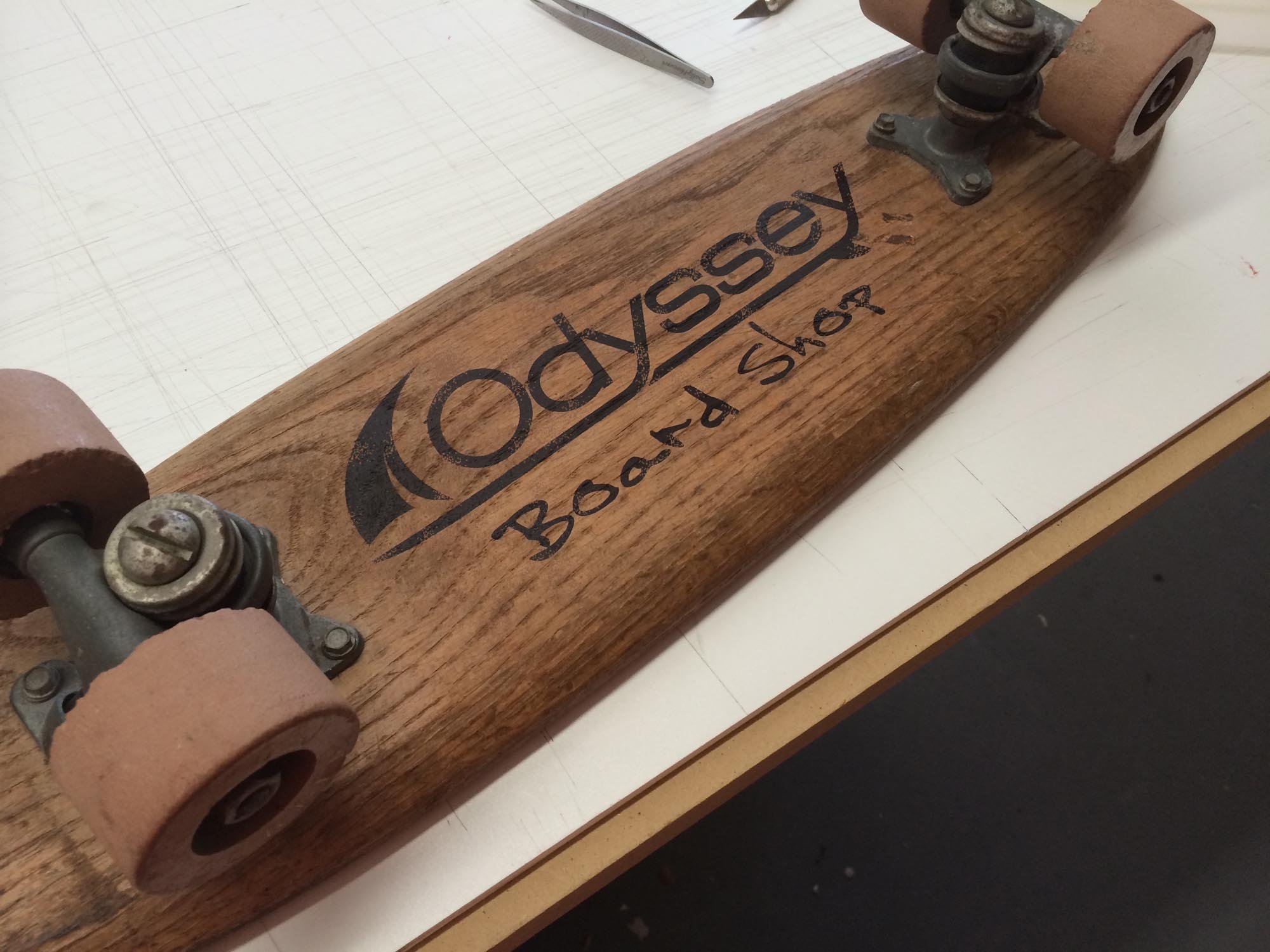 Close-up of a skateboard with a wooden deck labeled 'Board Shop' and 'Ossory,' showing pink wheels and metal trucks, on a white surface.