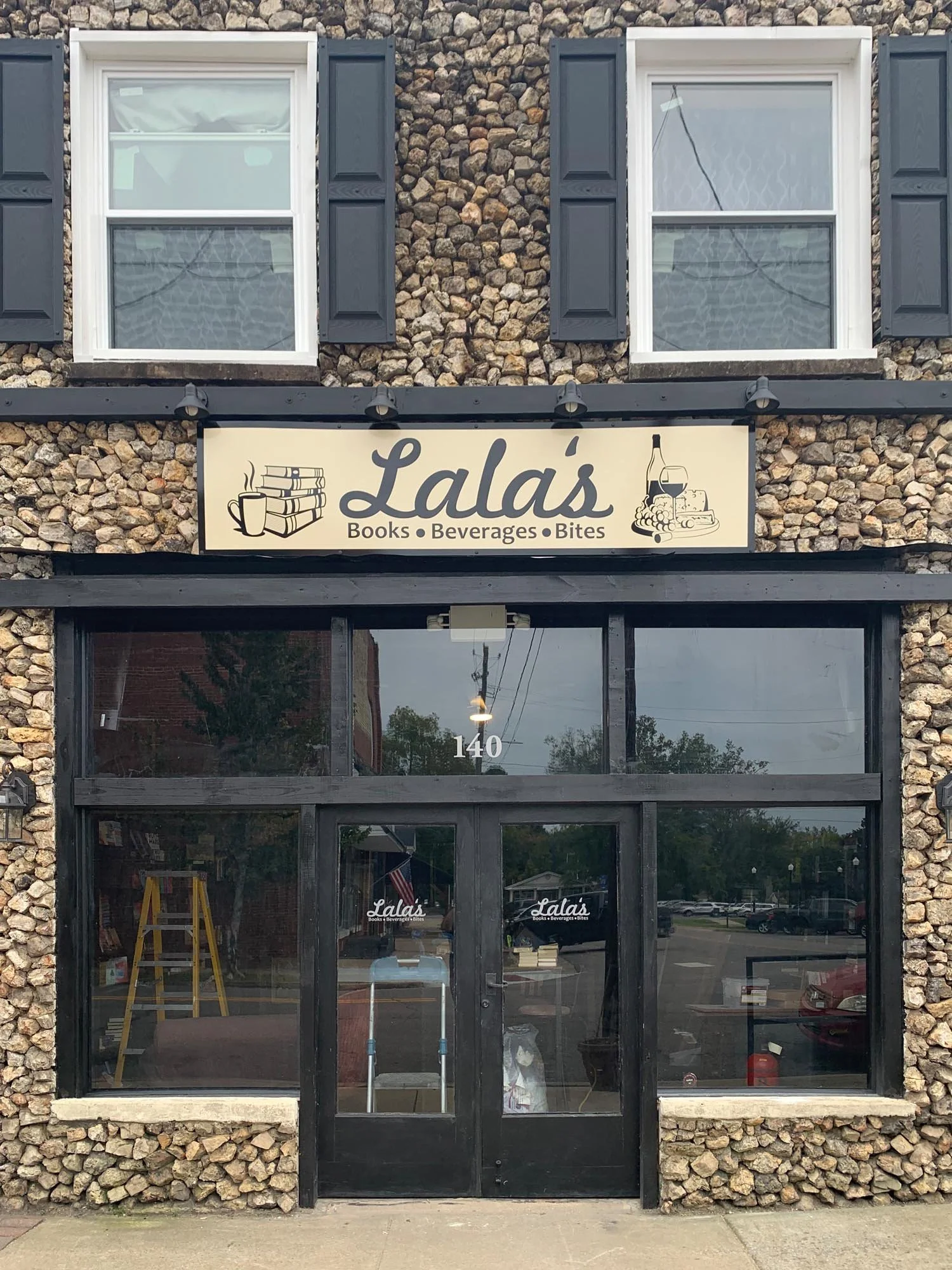 The storefront of Lala's, a shop that sells books, beverages, and bites. The building has a stone exterior, two windows with black shutters, and a black framed glass entrance door with the number 140. There are signs on the windows and above the door