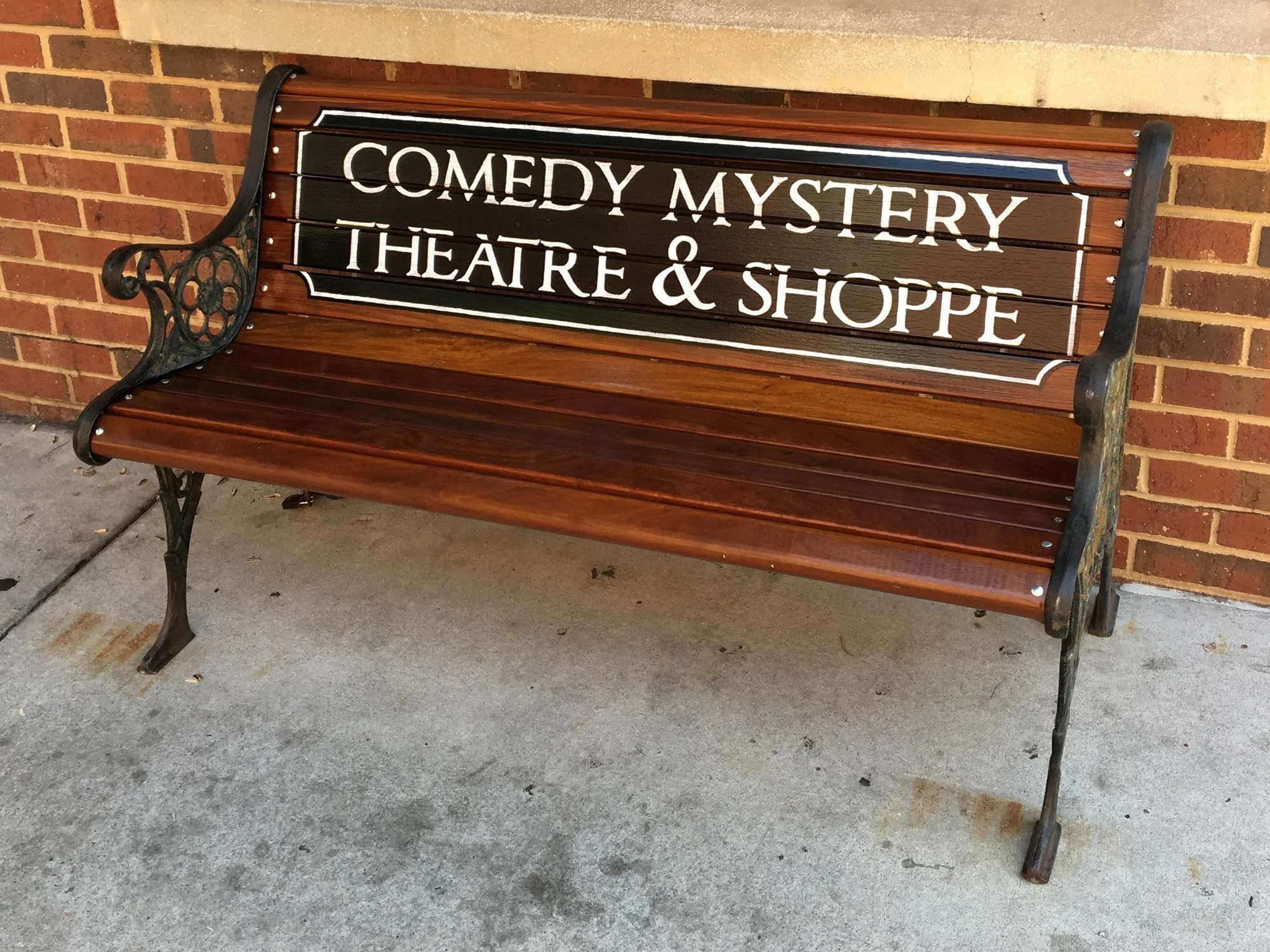A wooden and metal bench in front of a brick wall with a sign that reads 'Comedy Mystery Theatre & Shoppe'.