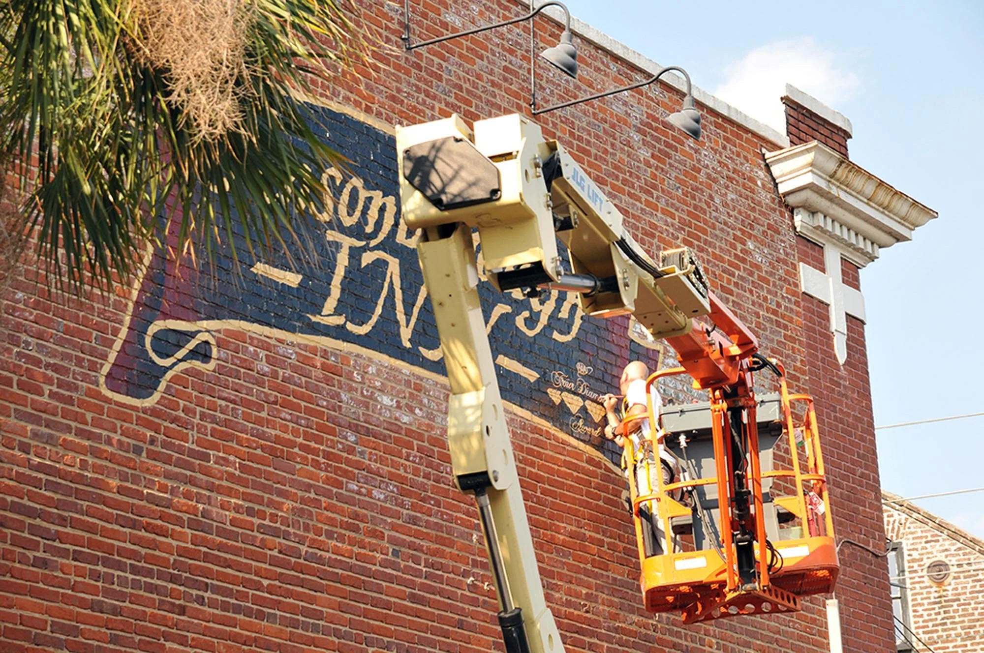 Two workers painting a mural on a brick building using an orange lift.
