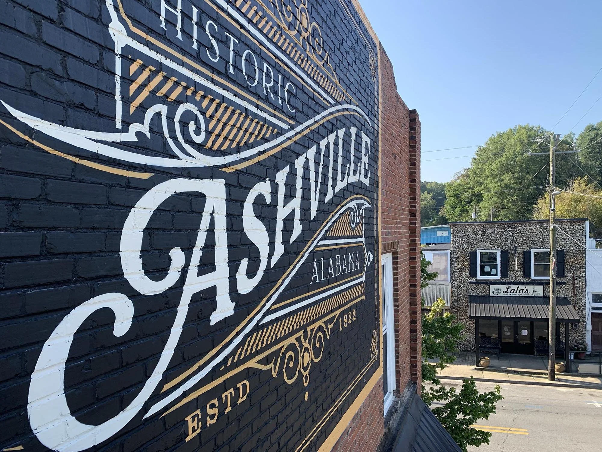 A painted sign on a brick building wall indicating 'Historic Cashion Alabama' with decorative scrollwork. In the background, there is a small stone building with a sign that reads 'Lala's,' and a street with utility poles and trees.