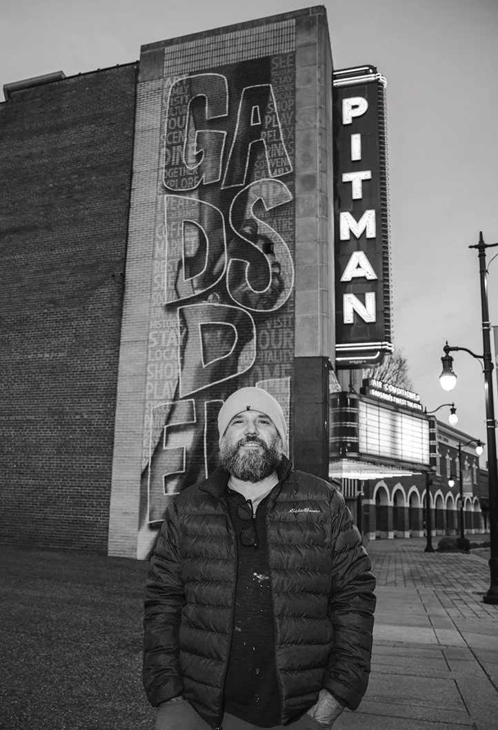 A man with a beard and beanie standing on a city sidewalk at night, smiling, in front of a theater with a large vertical illuminated sign reading 'PITMAN' and a large mural that says 'CADDYSHACK' on the building.