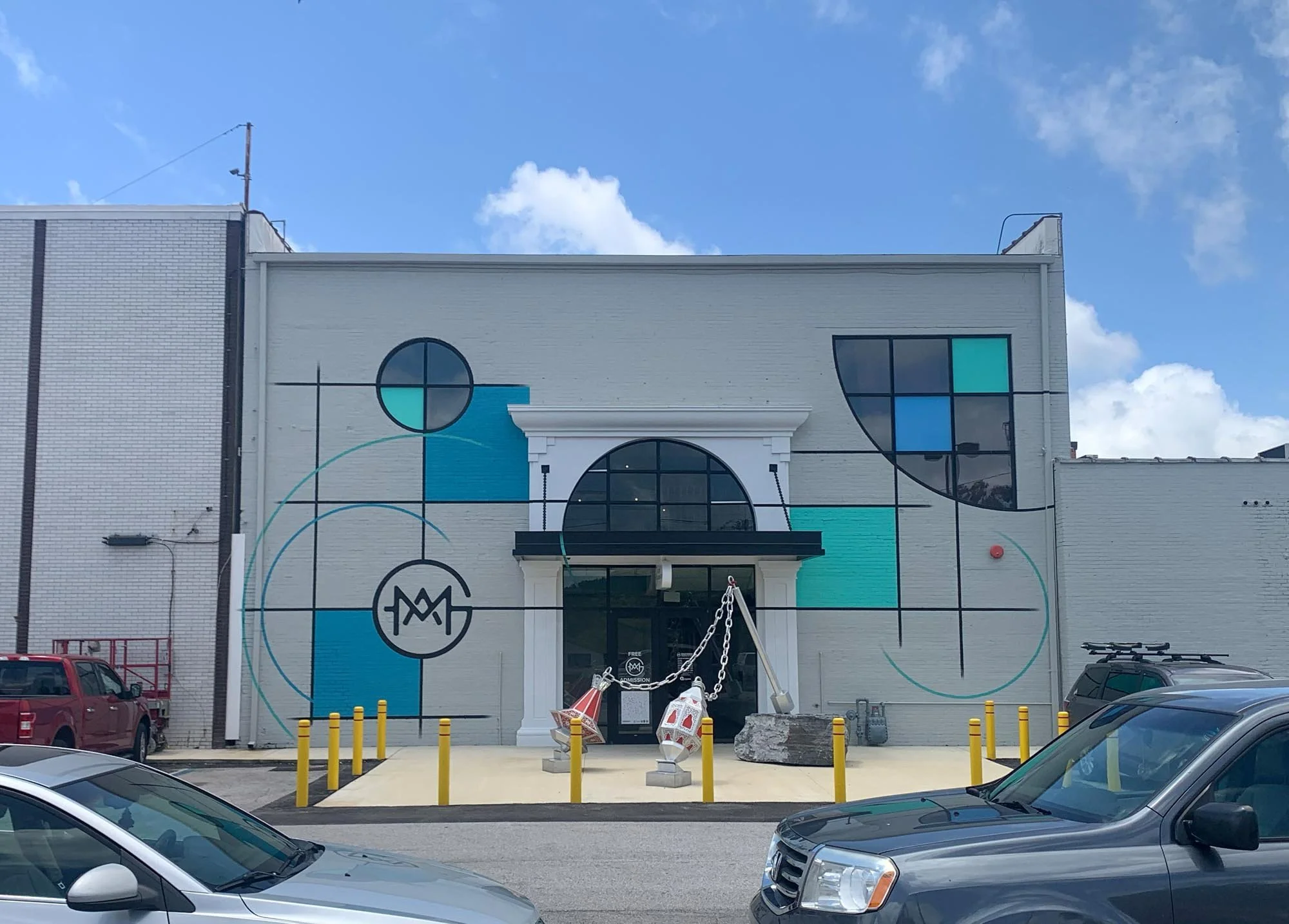 Colorful mural on a white building with abstract geometric shapes, circles, and lines in shades of black, blue, teal, and cyan, featuring a black circular logo with the initials 'GMA' in the center. There is a small entrance with a white surround, an