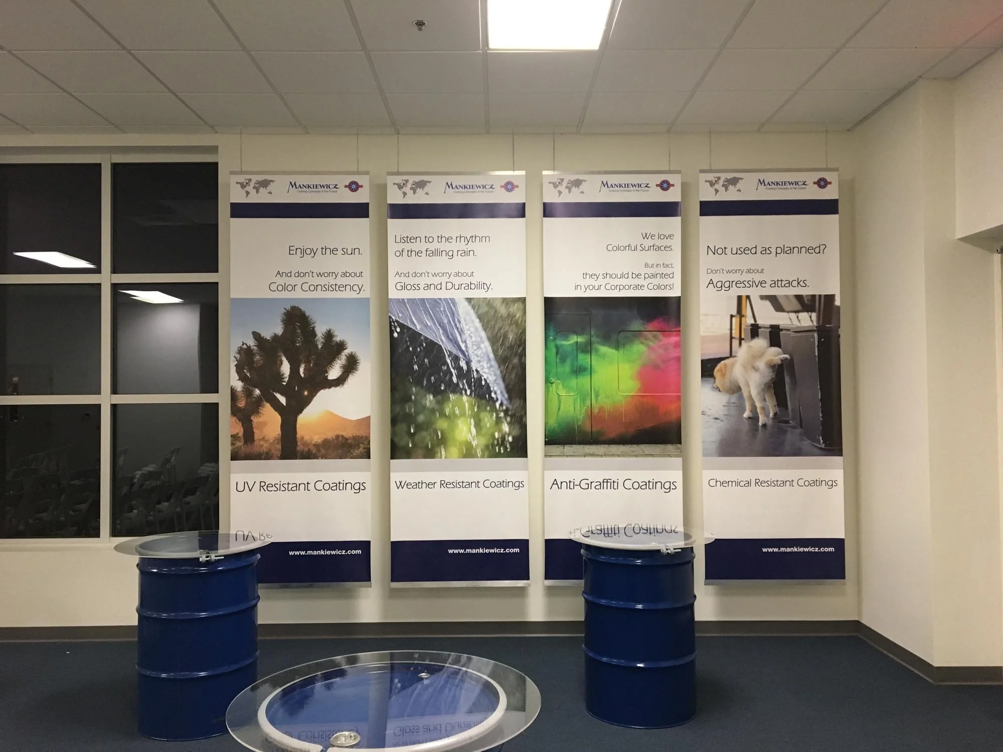 Indoor display with four vertical banners advertising different types of coatings: UV resistant, weather resistant, anti-graffiti, and chemical resistant. Each banner has a scenic or abstract image with descriptive text. There are two blue drums and 