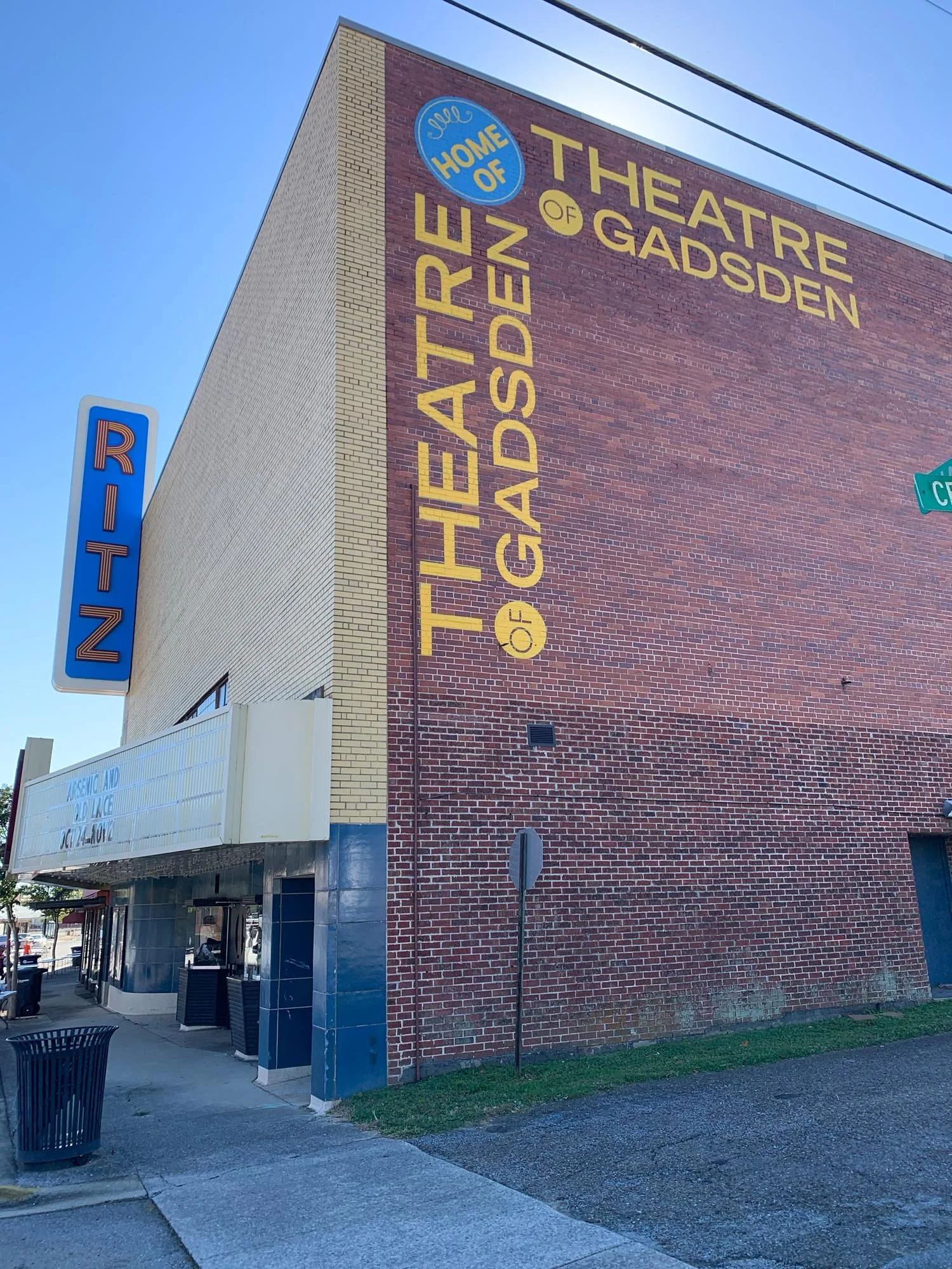 A brick building with a large painted sign reading 'Theatre of Gadsden, Home of Gadsden' on the side. There is a blue RITZ sign on the corner of the building and a street corner sign visible. The sky is clear and blue.