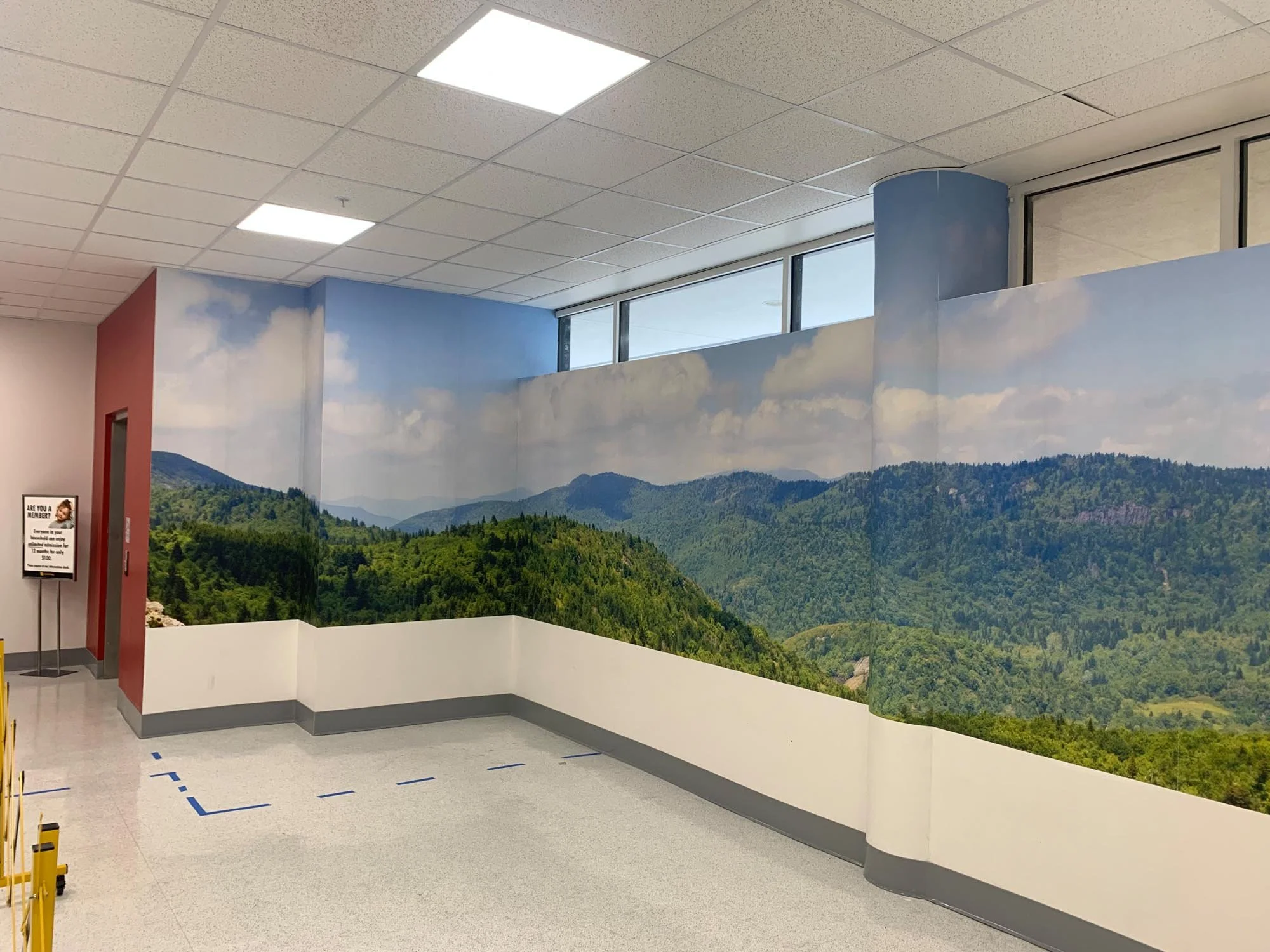 Indoor space with a wall mural of green mountains and blue sky, large windows, white ceiling, and gray flooring.
