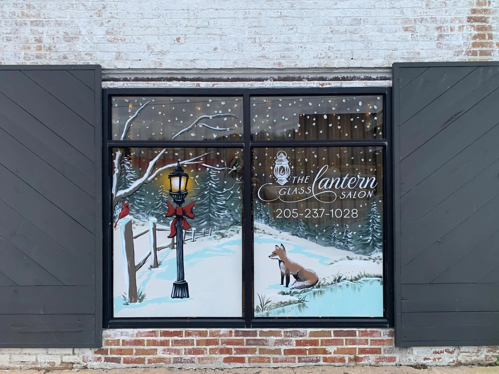 Window decal of a winter scene with snow-covered trees, a lamppost decorated with a red bow, and a fox sitting by a pond. Text on the window reads "The Lantern Glass Salon" along with a phone number.