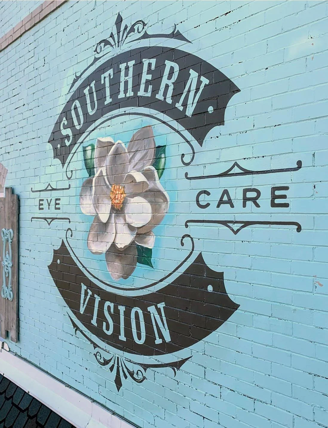 A mural on a light blue brick wall with the words 'Southern Vision' and 'Eye Care'. It features a white magnolia flower with green leaves surrounded by decorative black lines and scrolls.