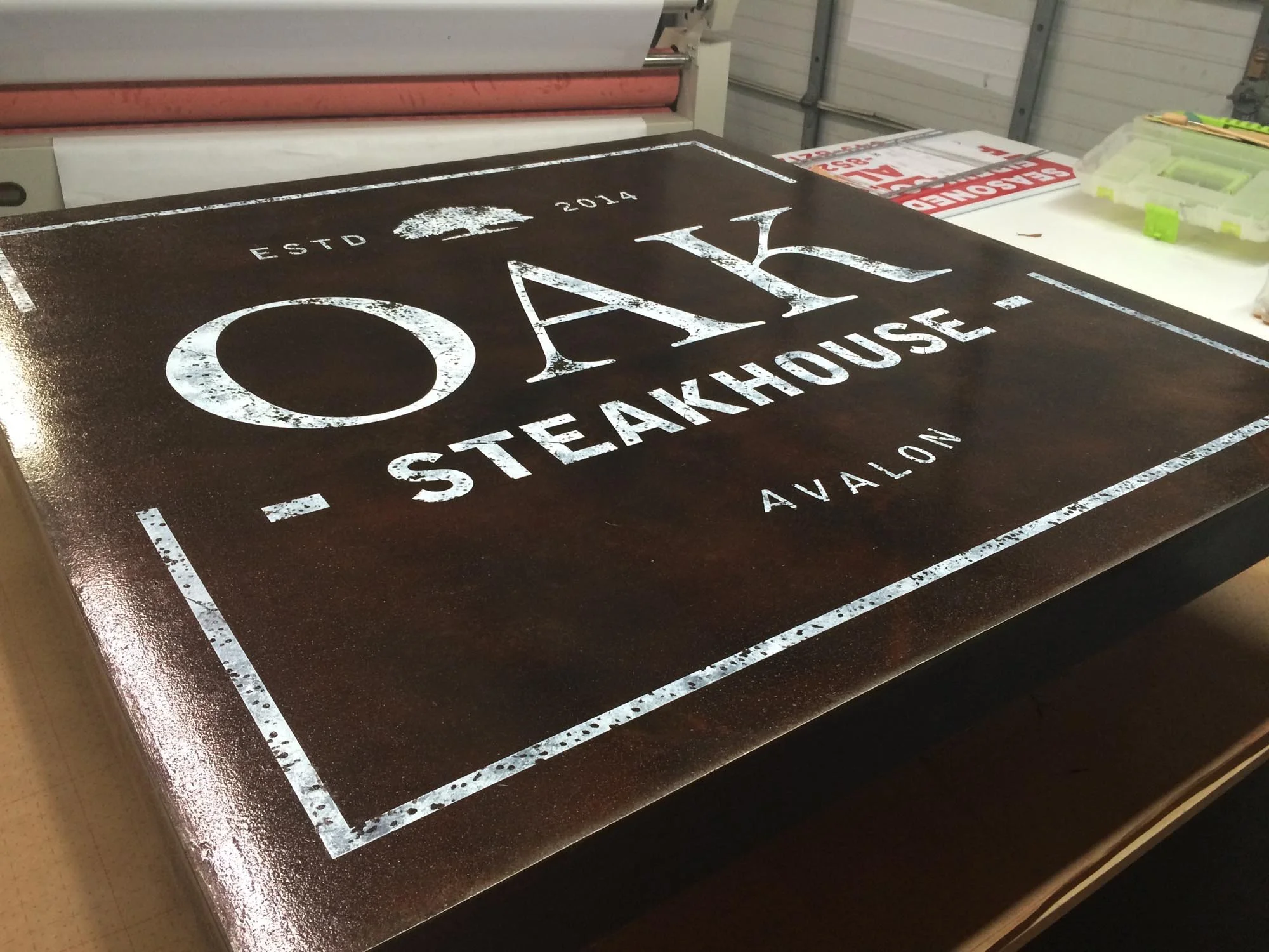 Close-up of a black and white sign for Oak Steakhouse with the text 'ESTD 2014' on top, the word 'OAK' in large letters in the middle, 'STEAKHOUSE' below it, and 'AVALON' at the bottom, placed on a table in a workshop or storage area.