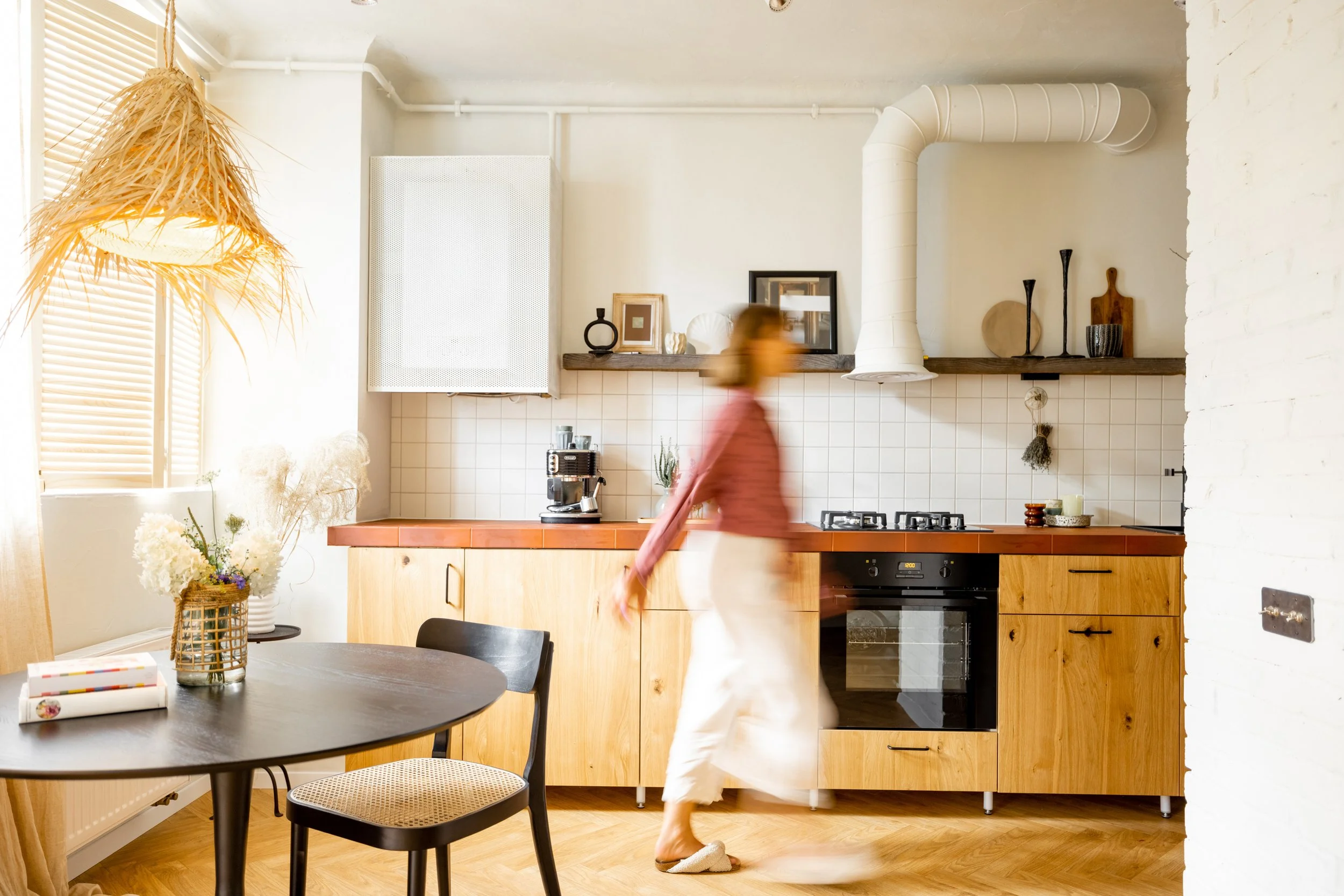 A woman walking past a kitchen with wooden cabinets, a black stove, and a white range hood. The kitchen has a wooden shelf with decorative items, and a table with a basket of flowers and some books. There is a large window with blinds on the left, and a woven light fixture hanging from the ceiling.