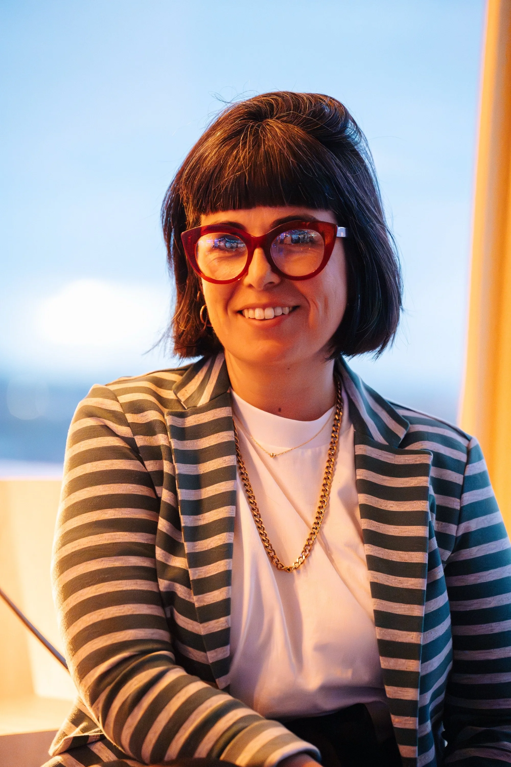 Woman smiling, wearing red glasses, striped blazer, white shirt, gold chains, indoors with blue background.