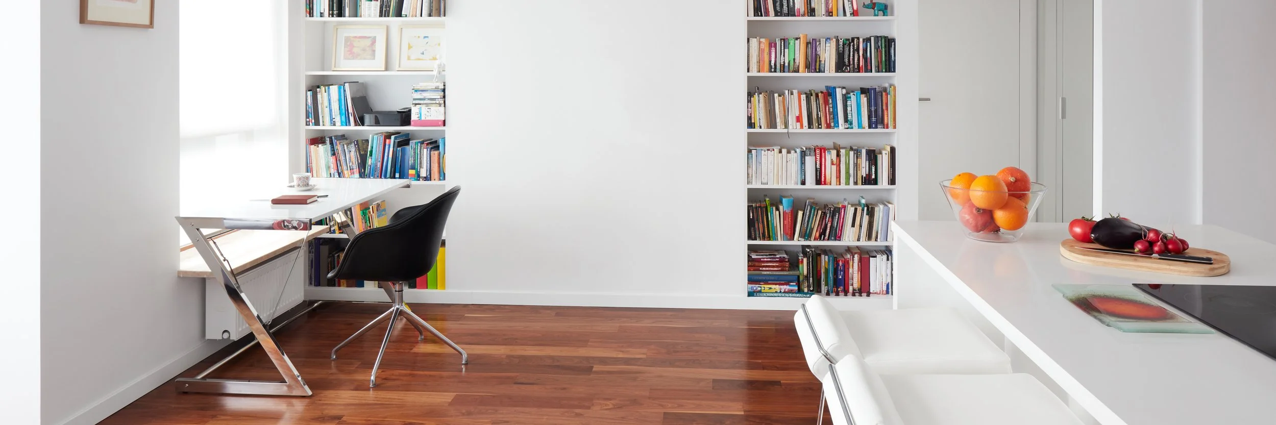 Modern white kitchen and library area with a counter, fruit bowl, and black chair.