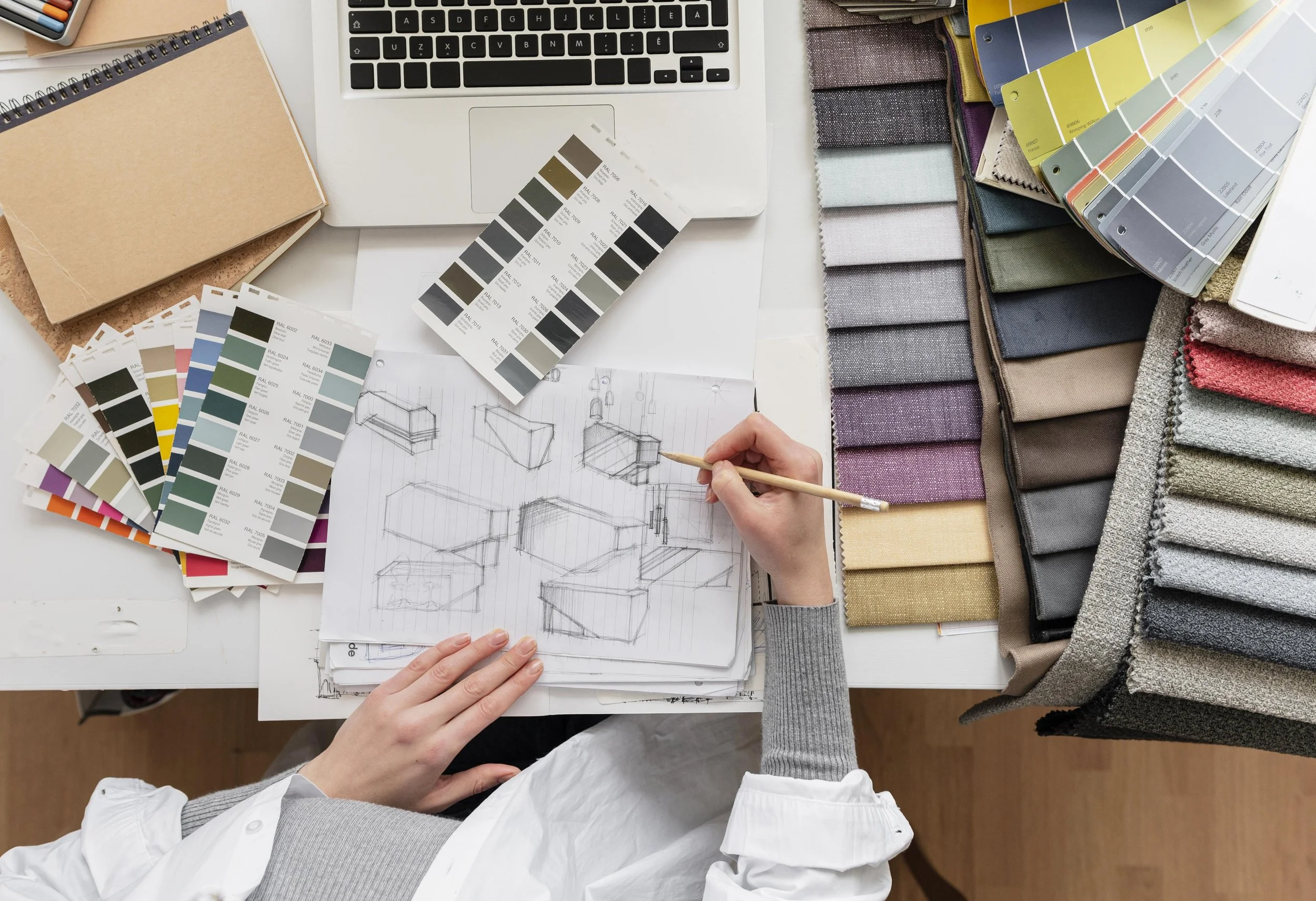 A person wearing a white shirt sketches furniture designs on lined paper, surrounded by color swatches and fabric samples, with a laptop nearby on a white desk.