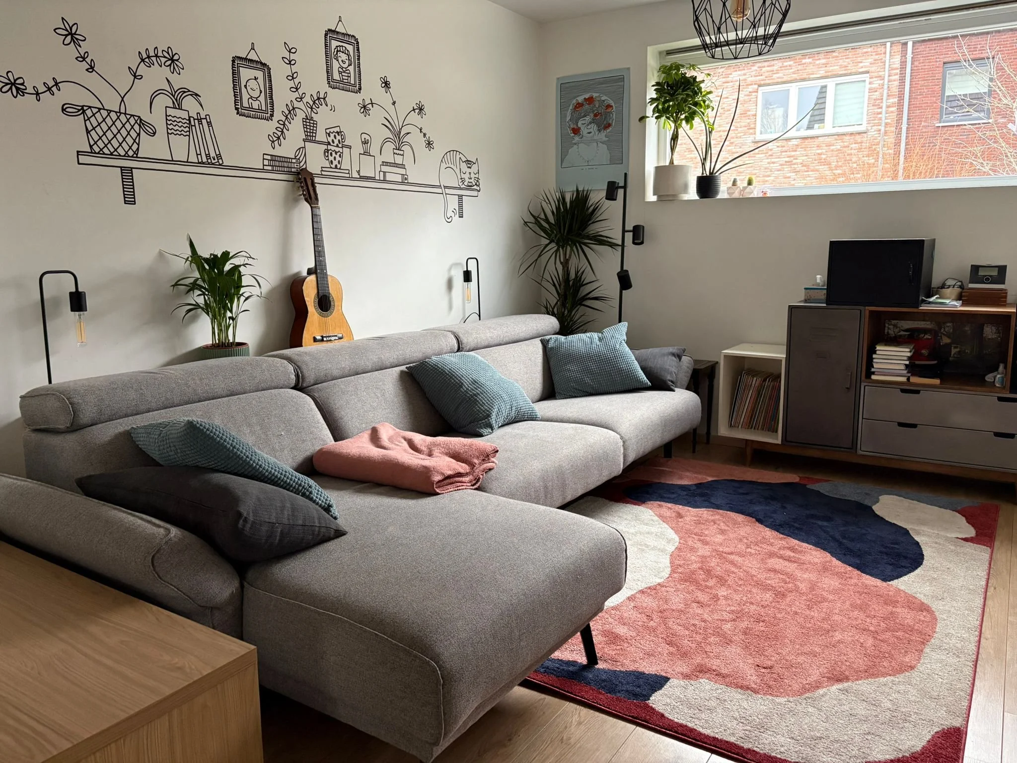Living room with a gray sofa, decorative pillows, a wooden side table, houseplants, wall art including a line drawing of a shelf with books and plants, a guitar, and a colorful abstract rug. Large window with view of brick building exterior.