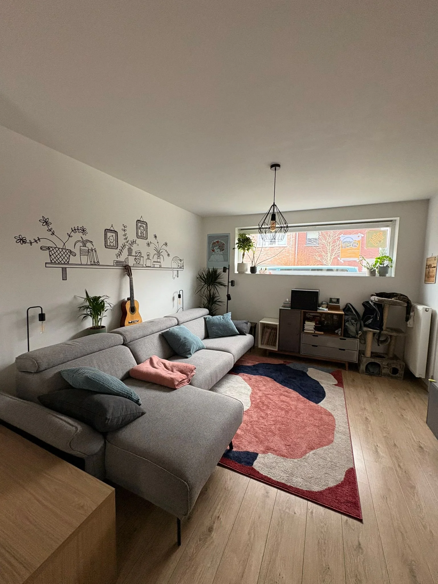 Cozy living room with a light gray fabric sofa decorated with pillows, a colorful abstract rug, a guitar leaning against the wall, a wooden side table, a black bookshelf, houseplants on the windowsill and next to the sofa, and a geometric wire pendan