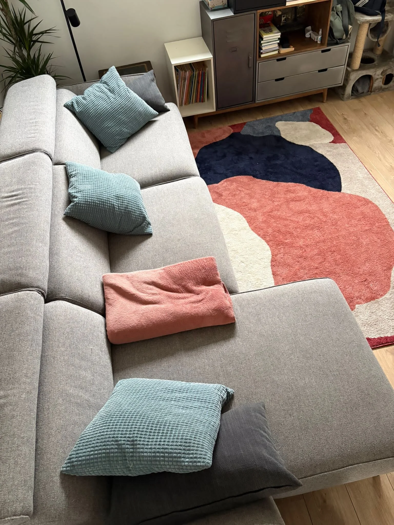 A beige sectional sofa with multiple textured pillows in shades of blue, gray, and pink, situated in a living room with a colorful abstract rug, a wooden floor, a bookcase, and a TV.