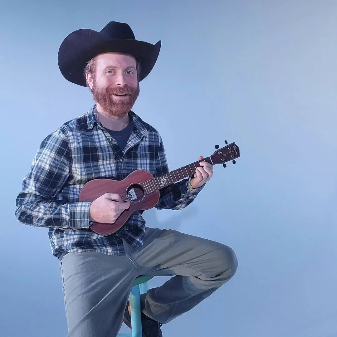 A man with red hair and beard wearing a large black cowboy hat, plaid shirt, and beige pants, sitting on a stool, playing a small ukulele, smiling against a plain blue background.