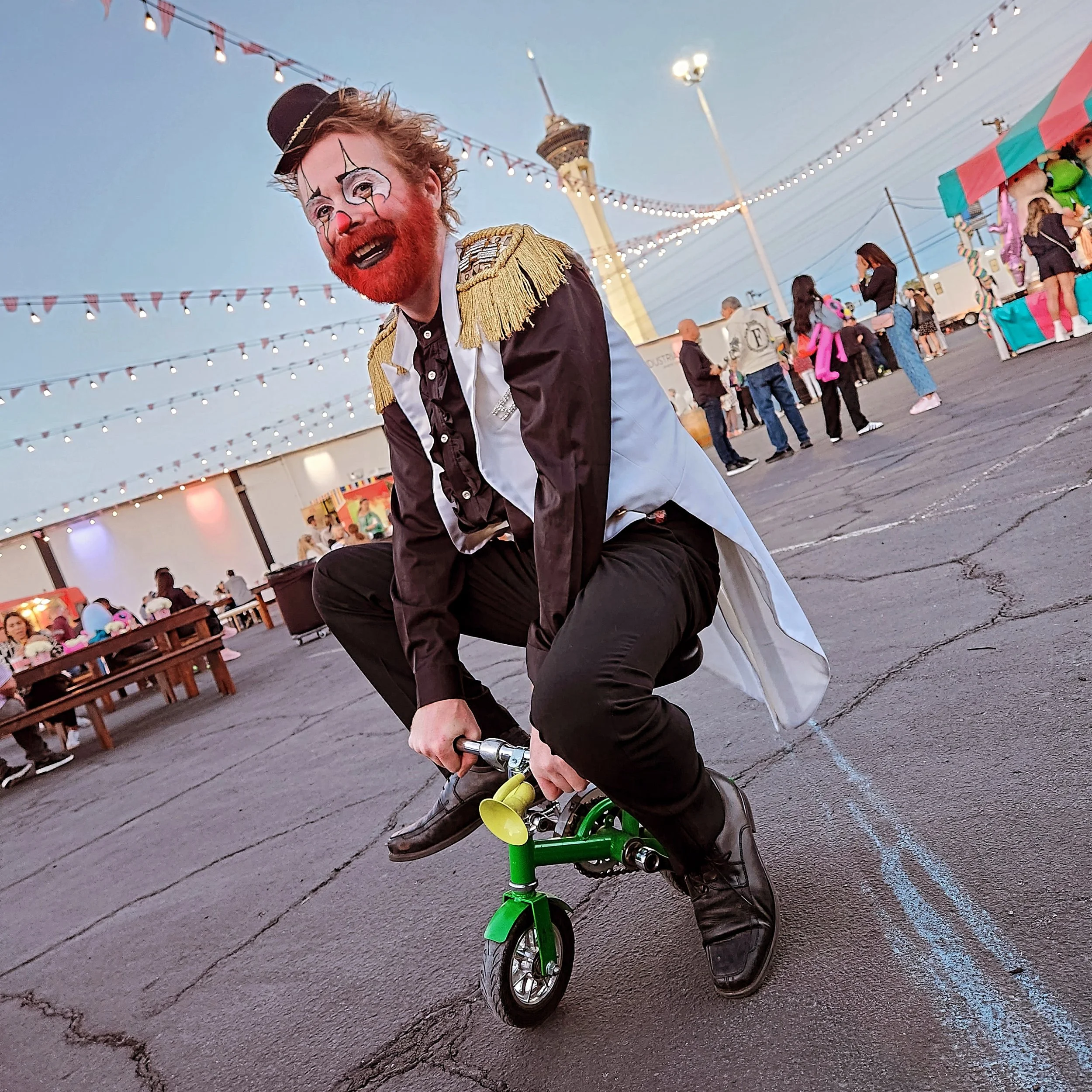 A clown with a painted face, red hair, and a red beard riding a small green bicycle at an outdoor fair during evening.