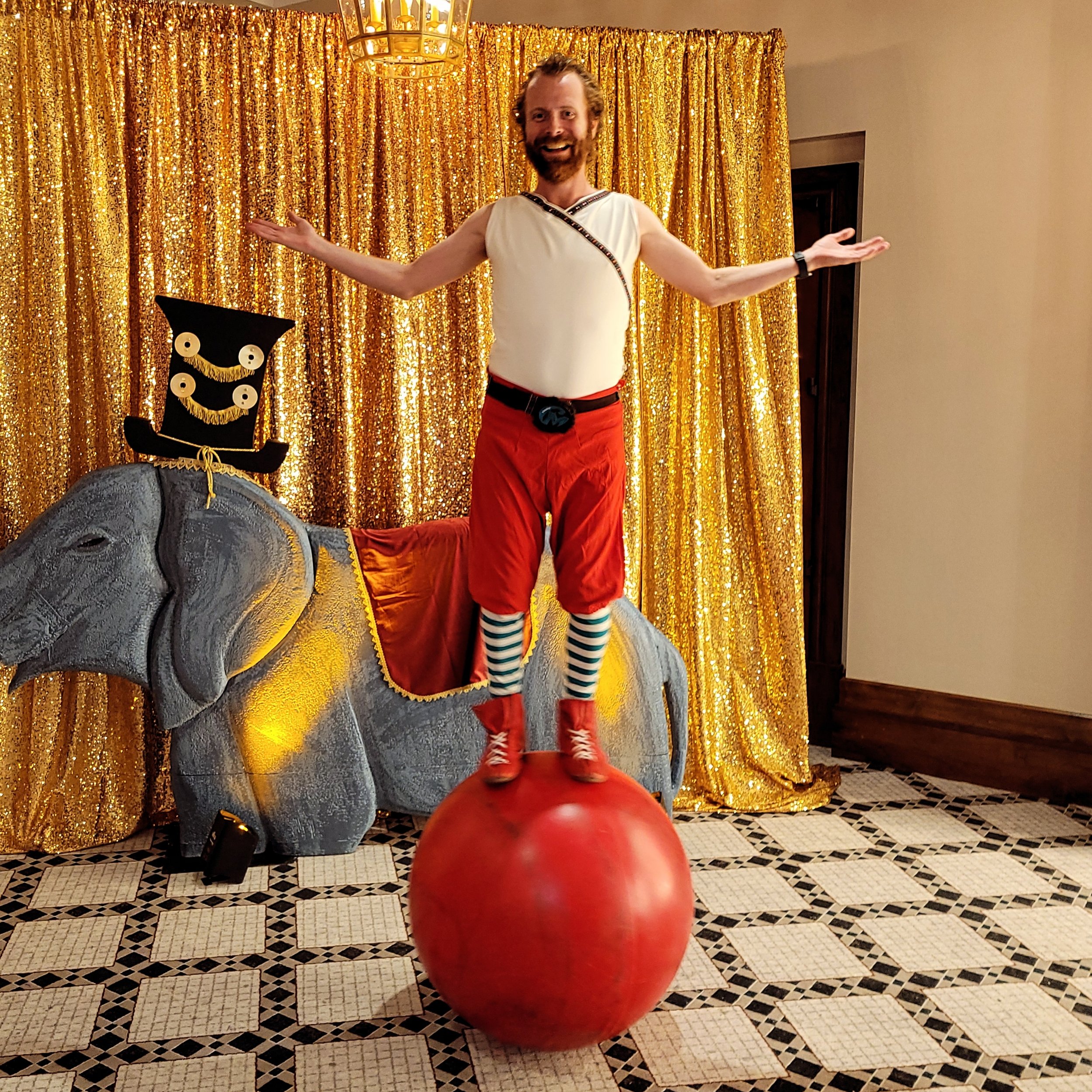 A man dressed as a circus performer standing with arms open on top of a large red ball in front of a golden curtain, with a decorated elephant statue and a smiling face mask on a stick beside him.