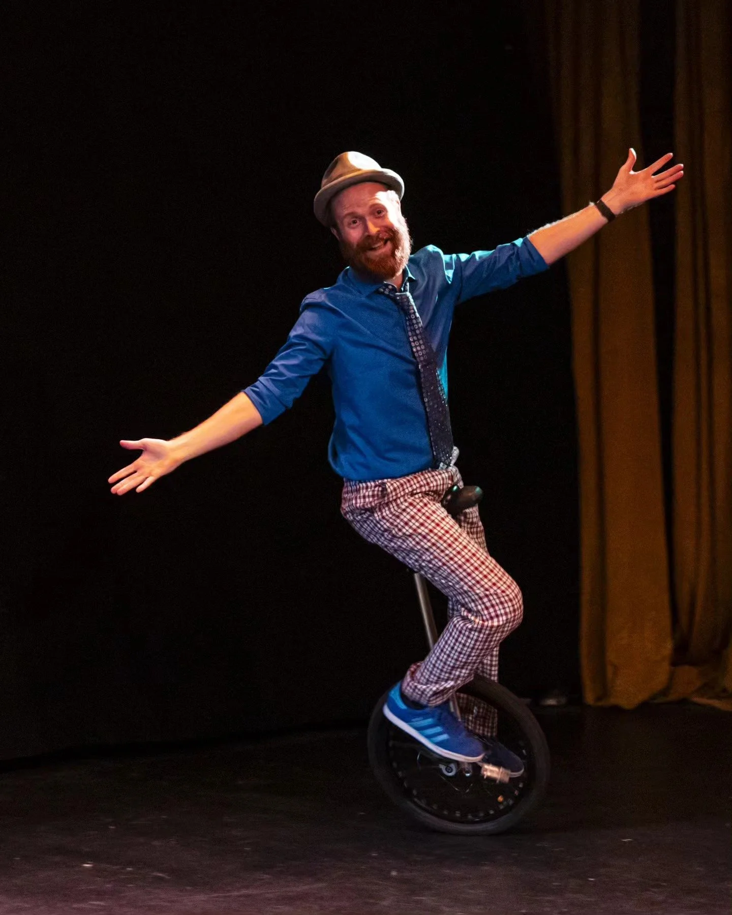 A man wearing a gray hat, blue shirt, patterned pants, and blue sneakers riding a unicycle on a stage, with arms spread wide.
