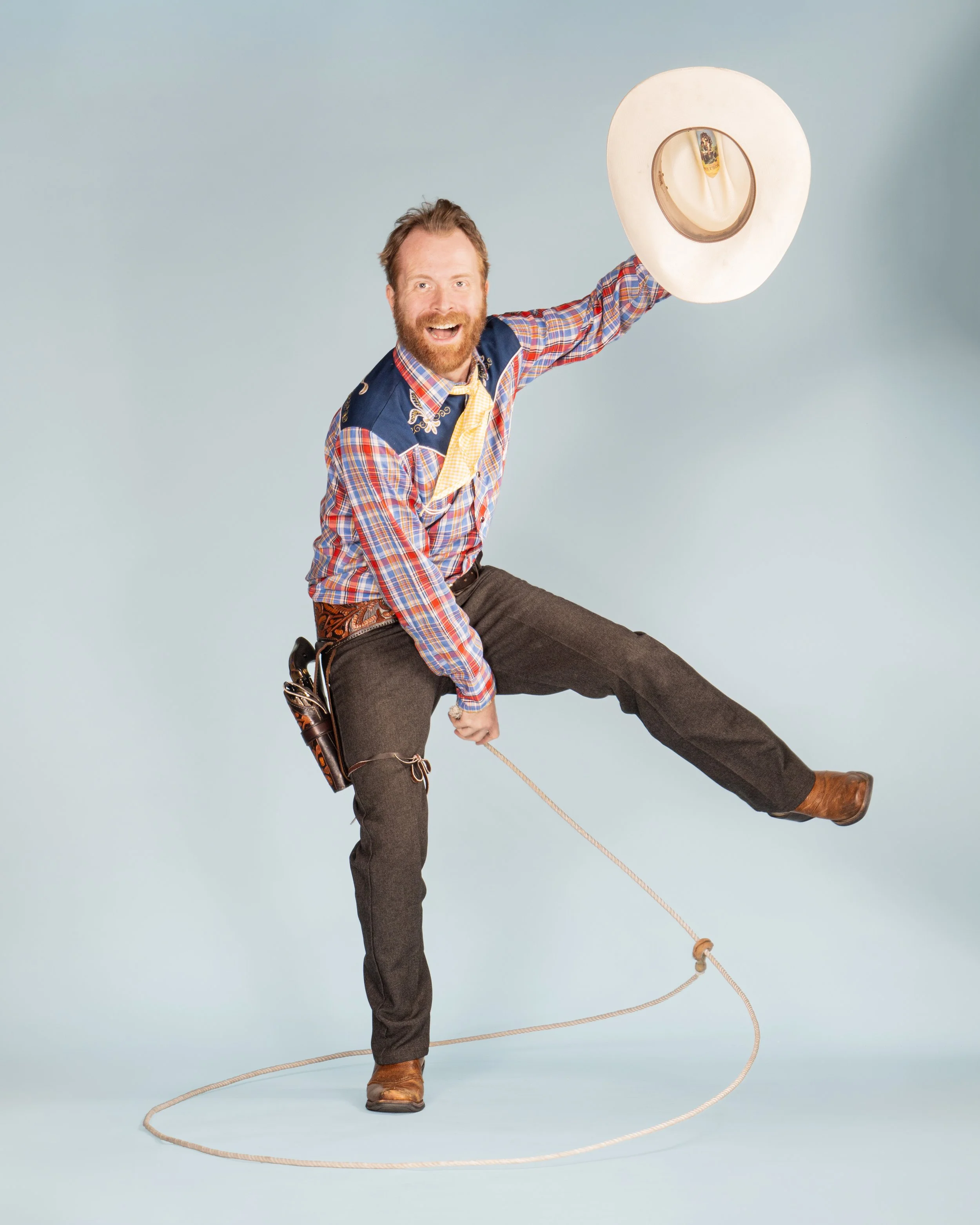 A cheerful man with a beard and reddish hair wearing Western attire, holding a cowboy hat, and lassoing with one leg raised, standing against a light blue background.