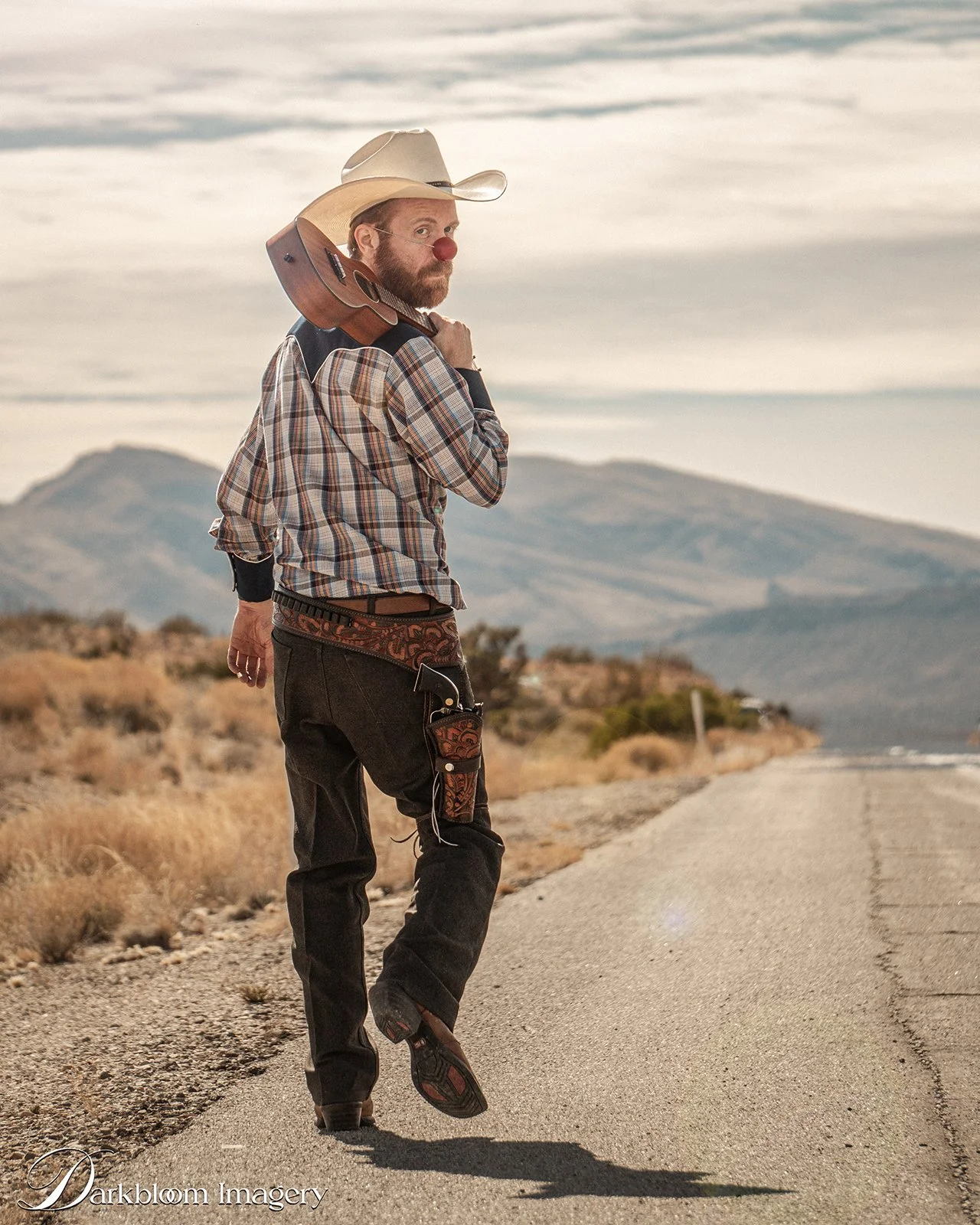 A man walking on a desert road at sunset, wearing a cowboy hat, plaid shirt, and carrying a guitar over his shoulder, with mountains in the background.
