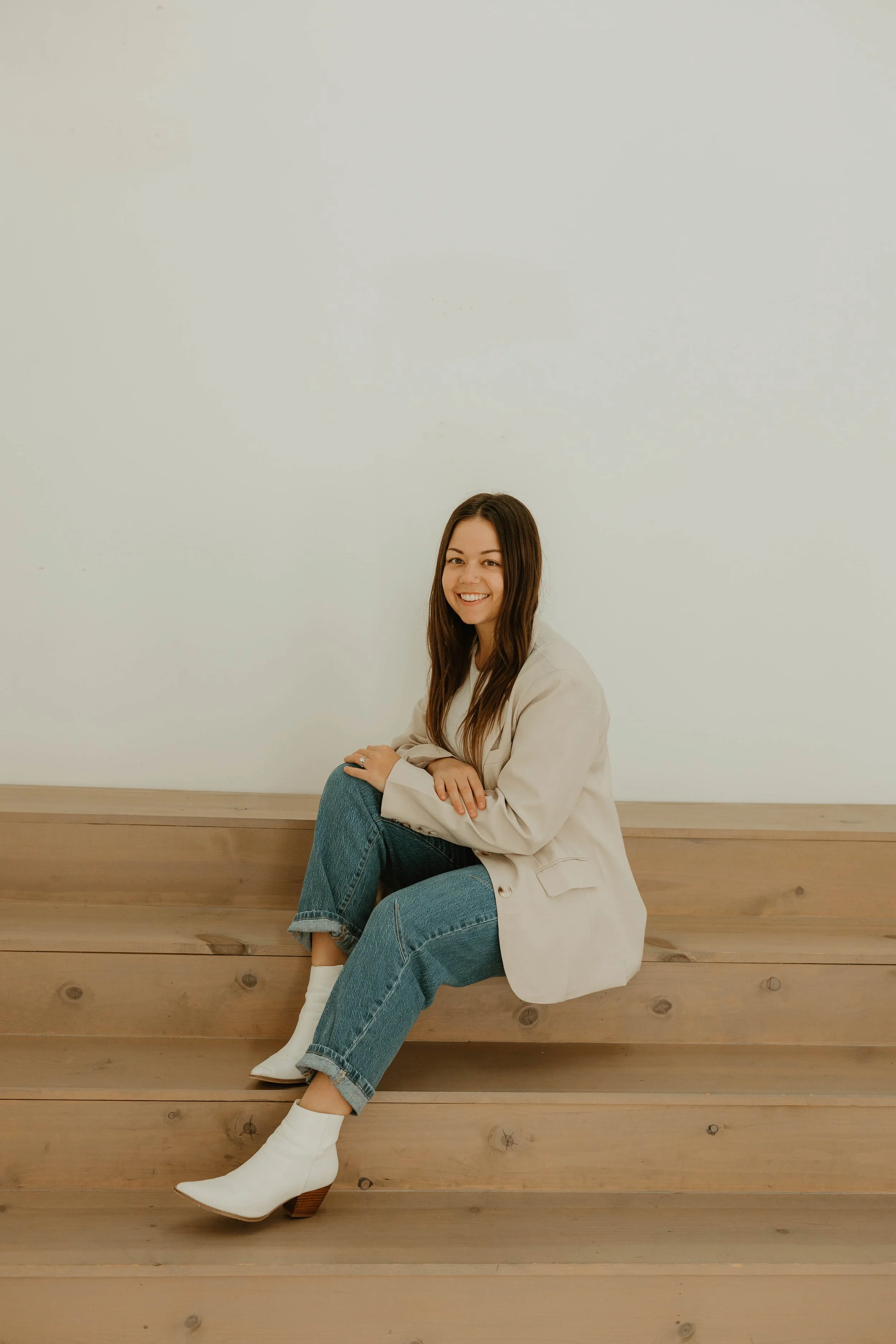 A young woman with long brown hair, wearing a beige blazer, blue jeans, and white ankle boots, sitting on wooden steps against a plain white wall, smiling.