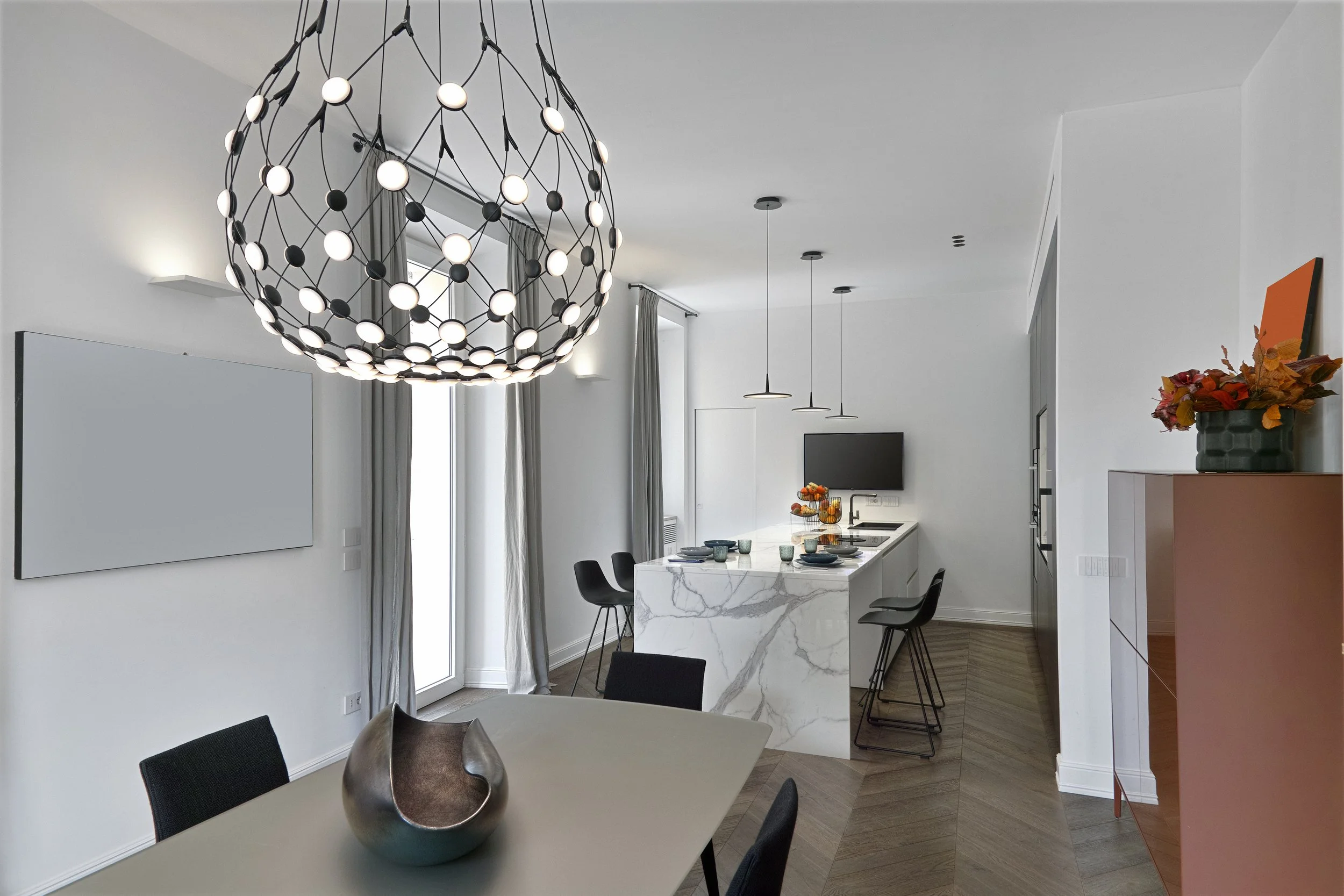 Modern kitchen and dining area with a large spherical pendant light, a marble kitchen island, black bar stools, and a wall-mounted TV. There are decorative items on the table and a vase with autumn leaves on a pink partition.