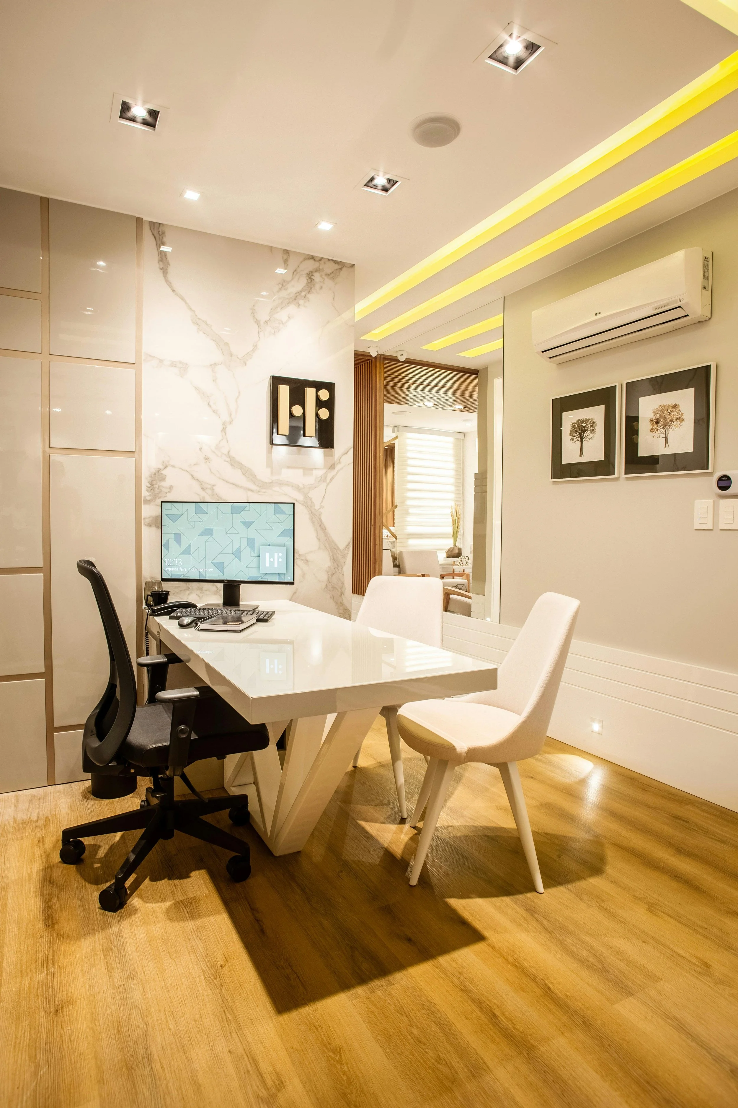Modern office with a white desk, black ergonomic chair, and two white chairs, against a marble wall with mounted artwork. A computer monitor, keyboard, and mouse are on the desk. The ceiling has recessed lighting and a yellow LED strip, and there is an air conditioning unit on the wall.