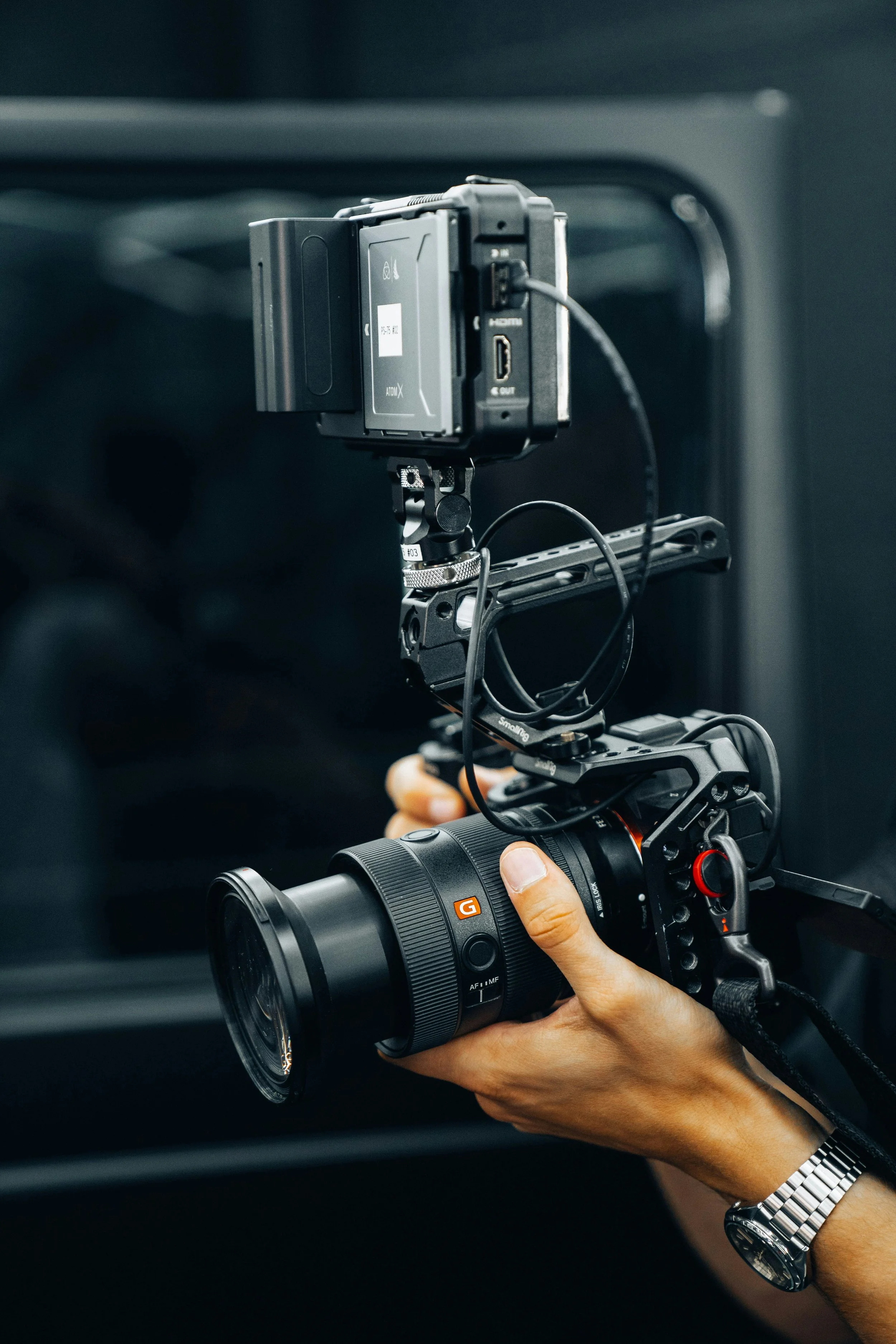 Close-up of a person holding a professional camera with a mounted external monitor, set against a dark background.