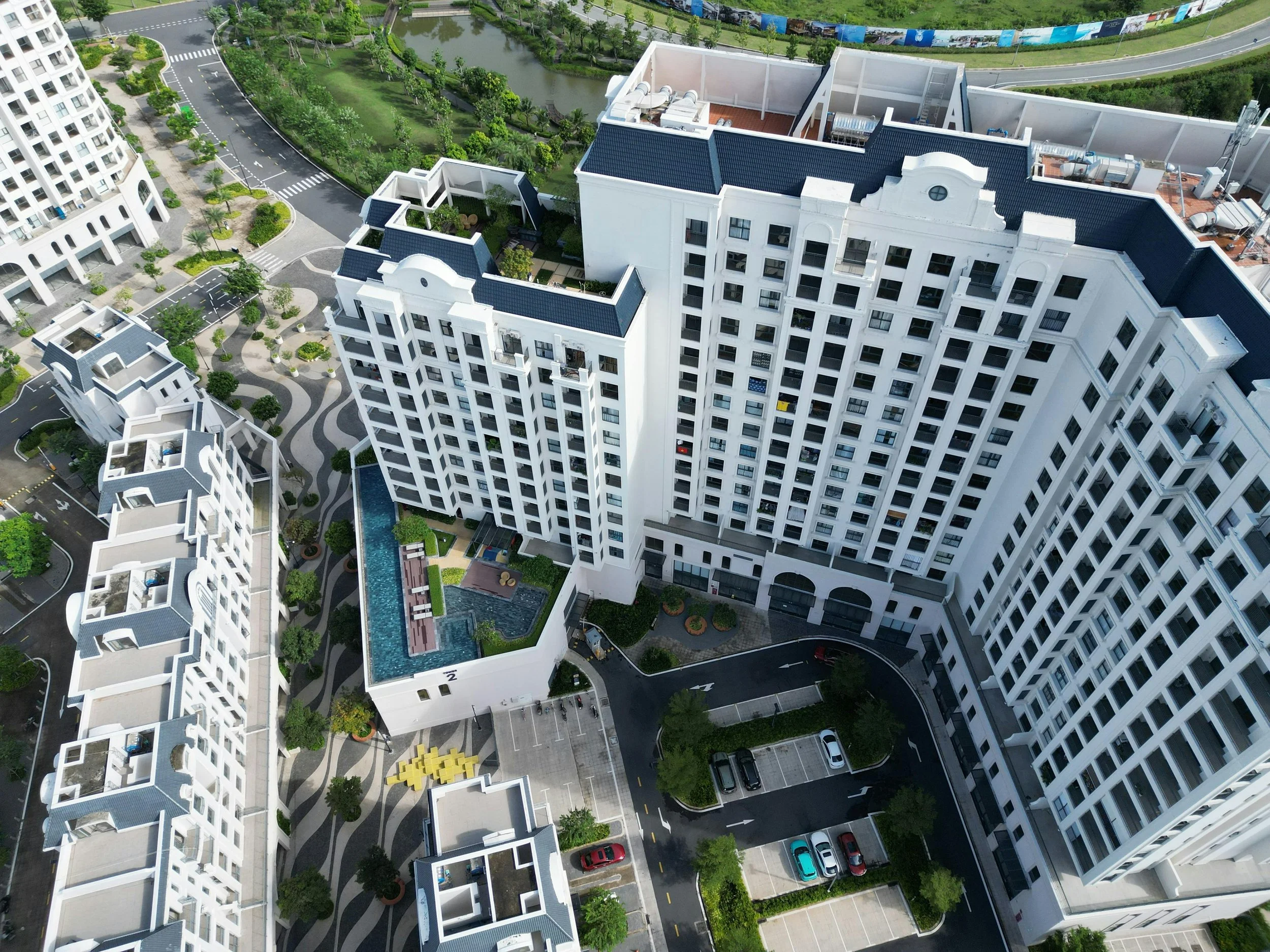 Aerial view of a modern white apartment complex with a swimming pool, parking lot, landscaped gardens, and adjacent roads.