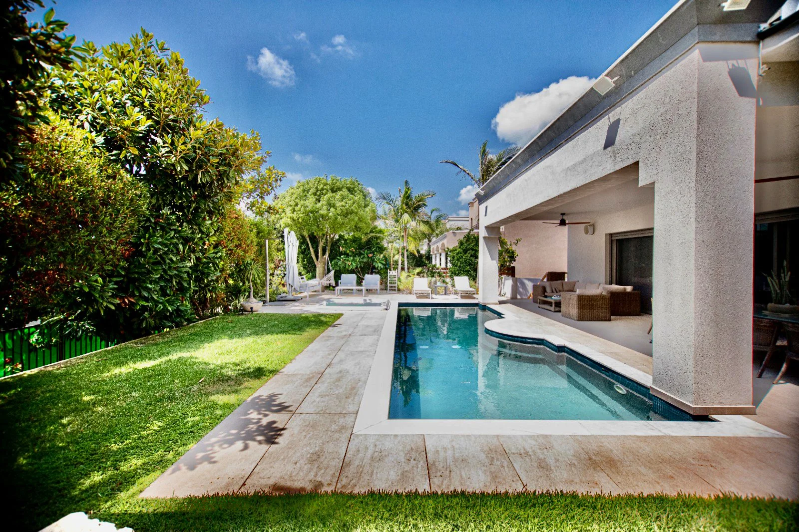 Swimming pool in backyard with outdoor furniture, lush green trees, and a white house under a partly cloudy blue sky.