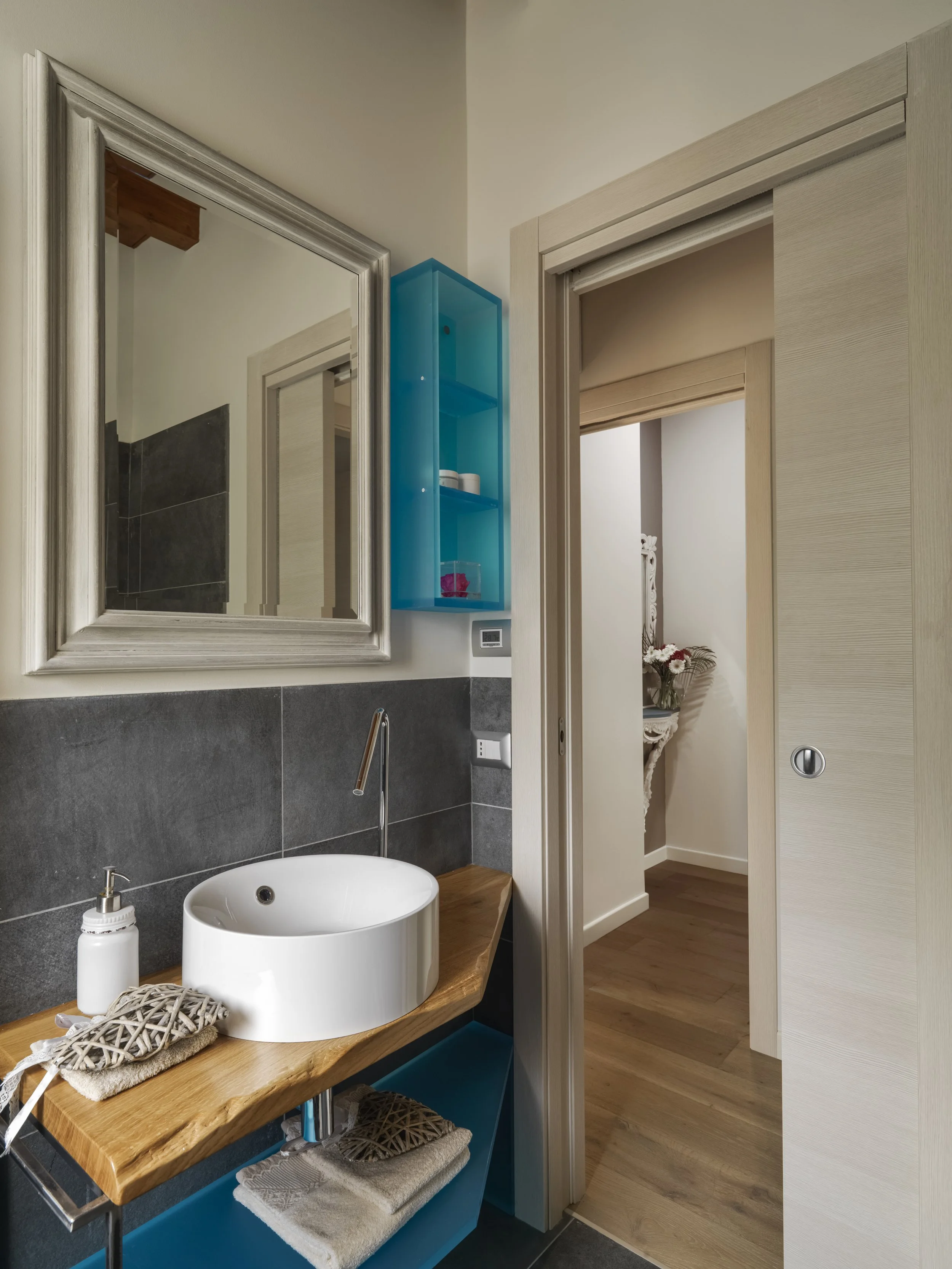 Modern bathroom with a white round vessel sink on a wooden countertop, a large silver-framed mirror, a small blue wall-mounted shelf, and a view into another room with a decorative flower arrangement.