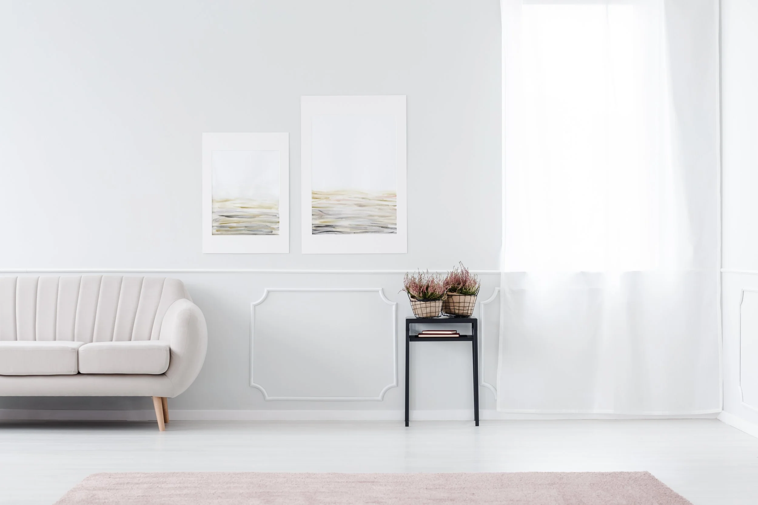 Minimalist living room with a white sofa, two framed abstract paintings on the wall, a small black side table with baskets of pink flowers, sheer white curtains, and a blush-colored rug.