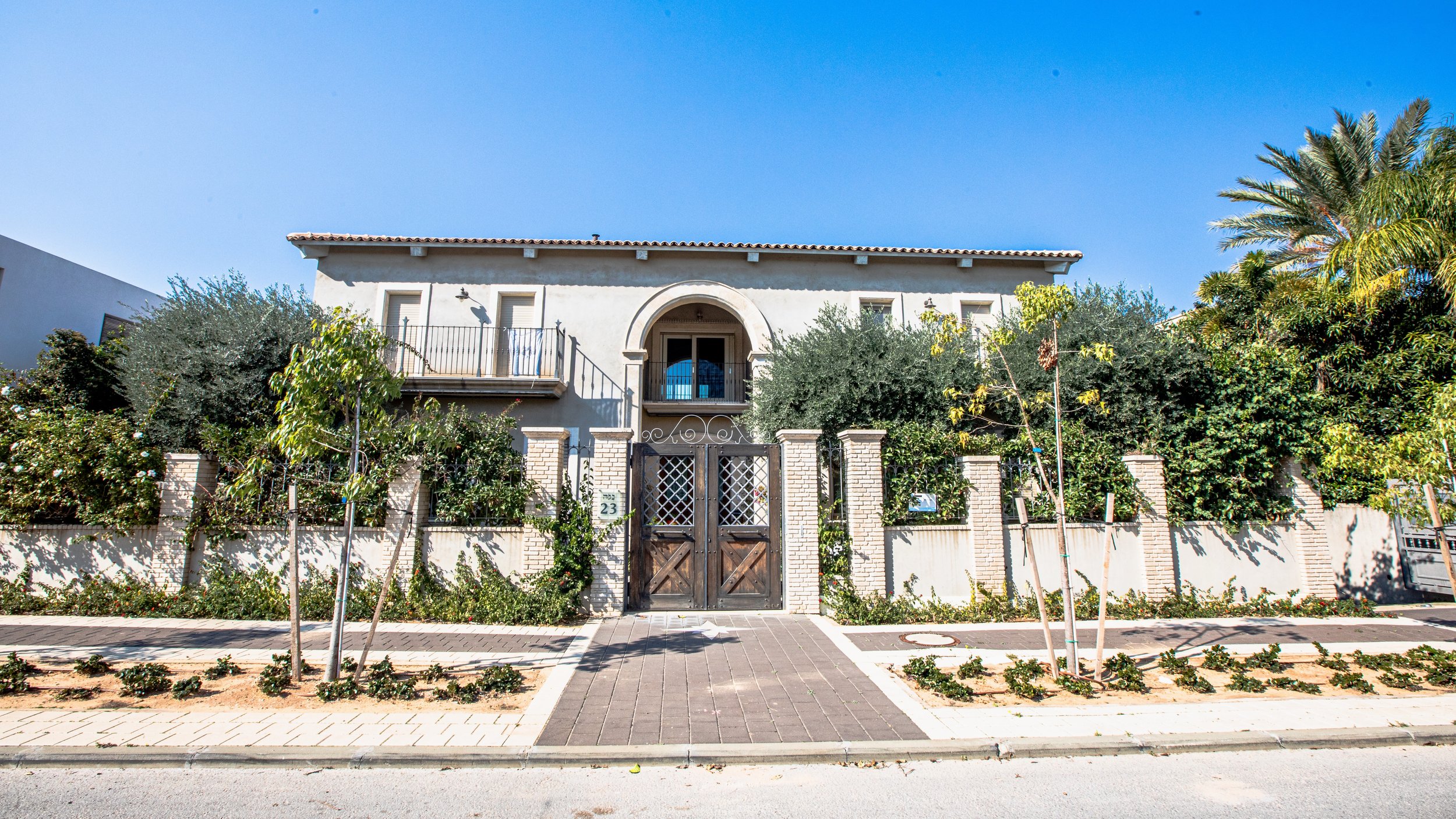 White two-story house with a tile roof, surrounded by trees, bushes, and a brick fence with a wooden gate, on a sunny day.