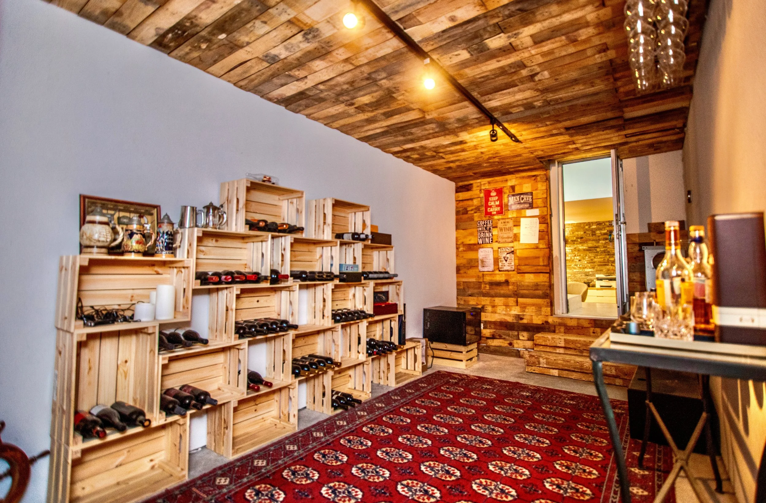 Interior of a room with a wooden ceiling, white walls, and a wooden accent wall. There are wooden crates arranged as shelves holding wine bottles, candles, and decorative items. A red patterned rug covers the floor. A small black speaker is on the fl