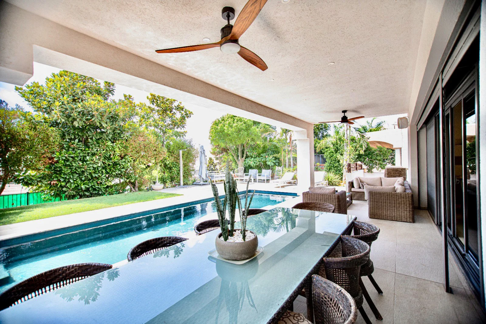 Covered outdoor patio area with a glass-top dining table, wicker chairs, and ceiling fans, overlooking a swimming pool and lush green trees.
