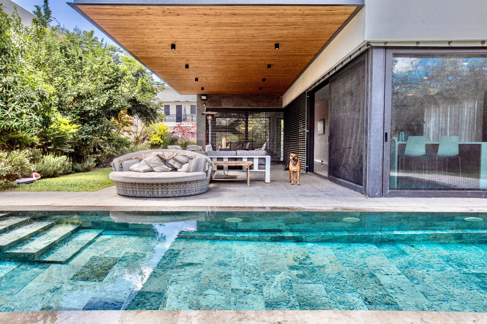 Modern house with a covered patio, outdoor seating area with cushions, trees and bushes in the background, clear swimming pool with steps leading in, and a dog standing on the patio.