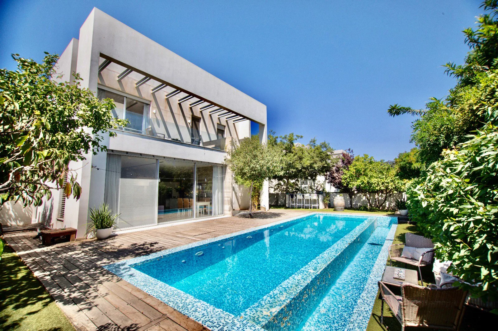 Modern house with large sliding glass doors, backyard swimming pool, patio chairs, umbrella, and lush green trees under a clear blue sky.