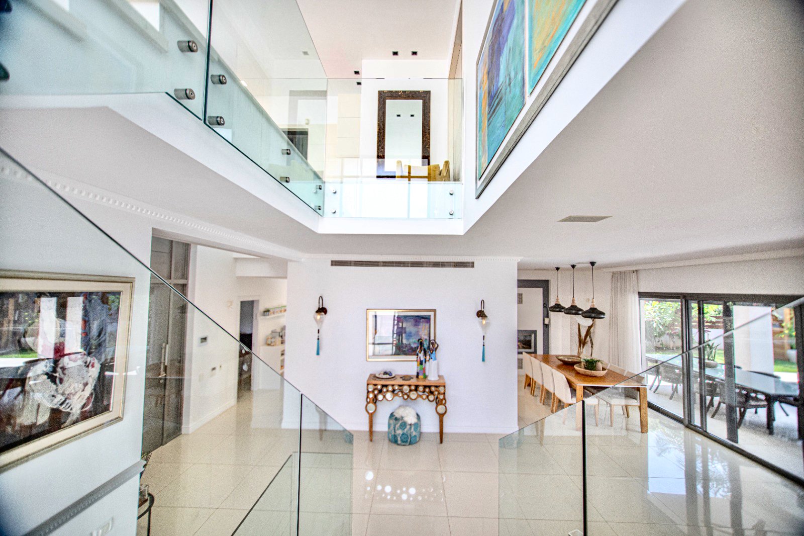 Interior view of a modern, airy living space with white walls, glass railing on the upper level, colorful abstract paintings, a wooden dining table with chairs, and patio doors leading outside.