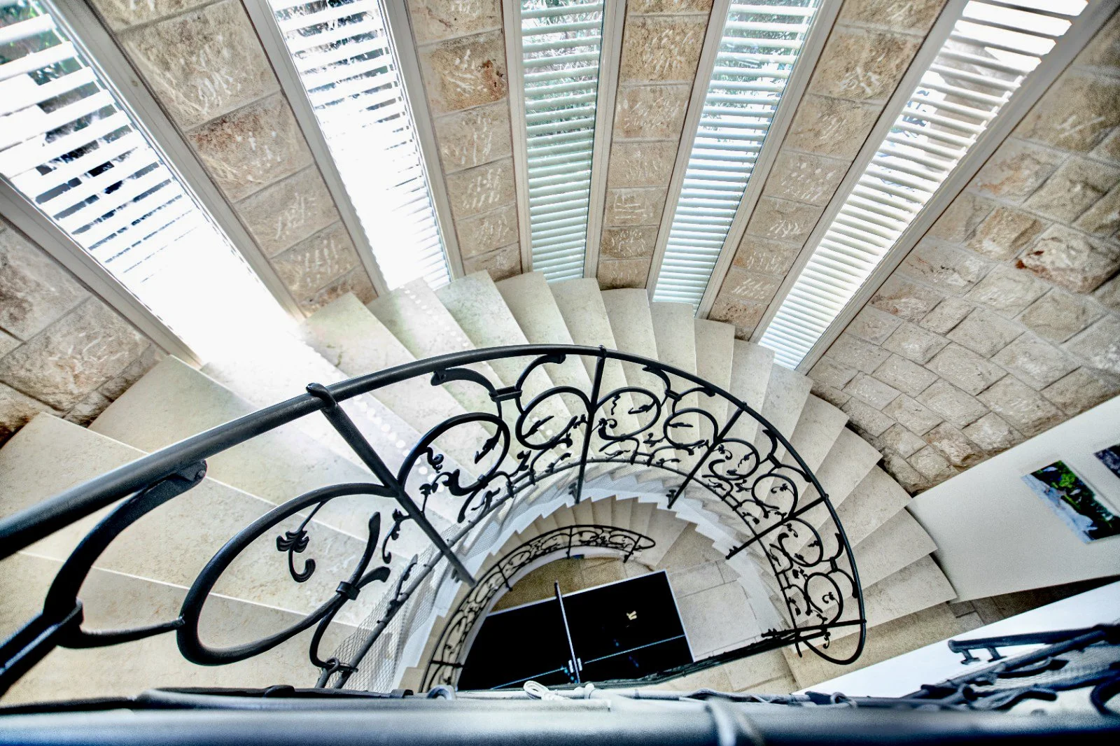 Looking down a spiral staircase with beige steps, a black ornate wrought iron railing, frontal window blinds, and a partial view of a stone wall.