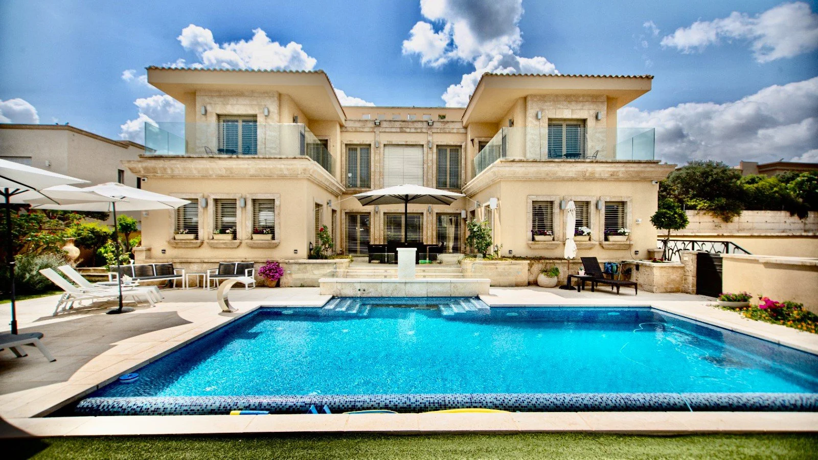 Luxury house with a swimming pool, outdoor seating, umbrellas, and landscaped garden under a partly cloudy sky.