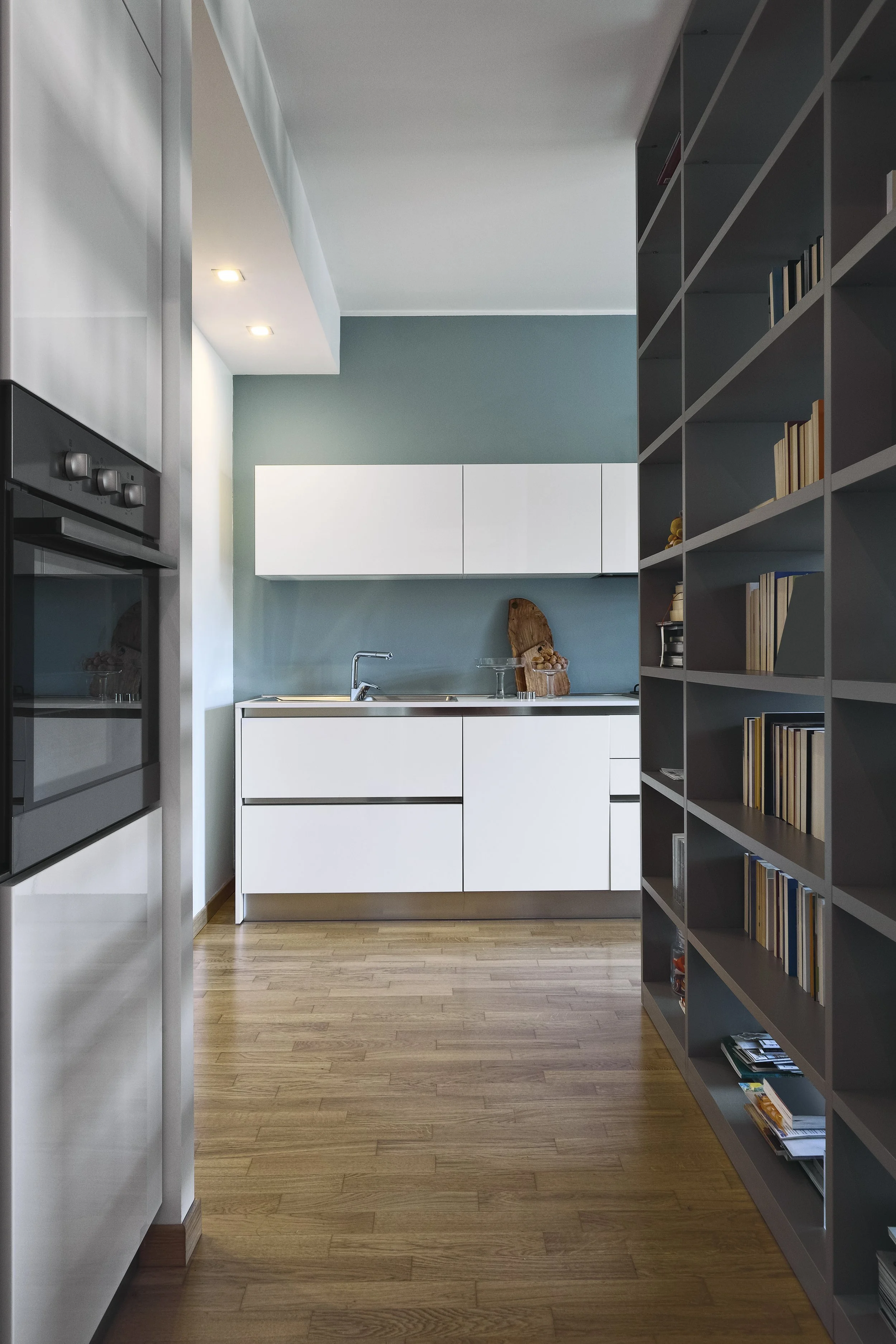 Modern kitchen with white cabinets, a blue wall, wooden floor, and a bookshelf filled with books on the right side.